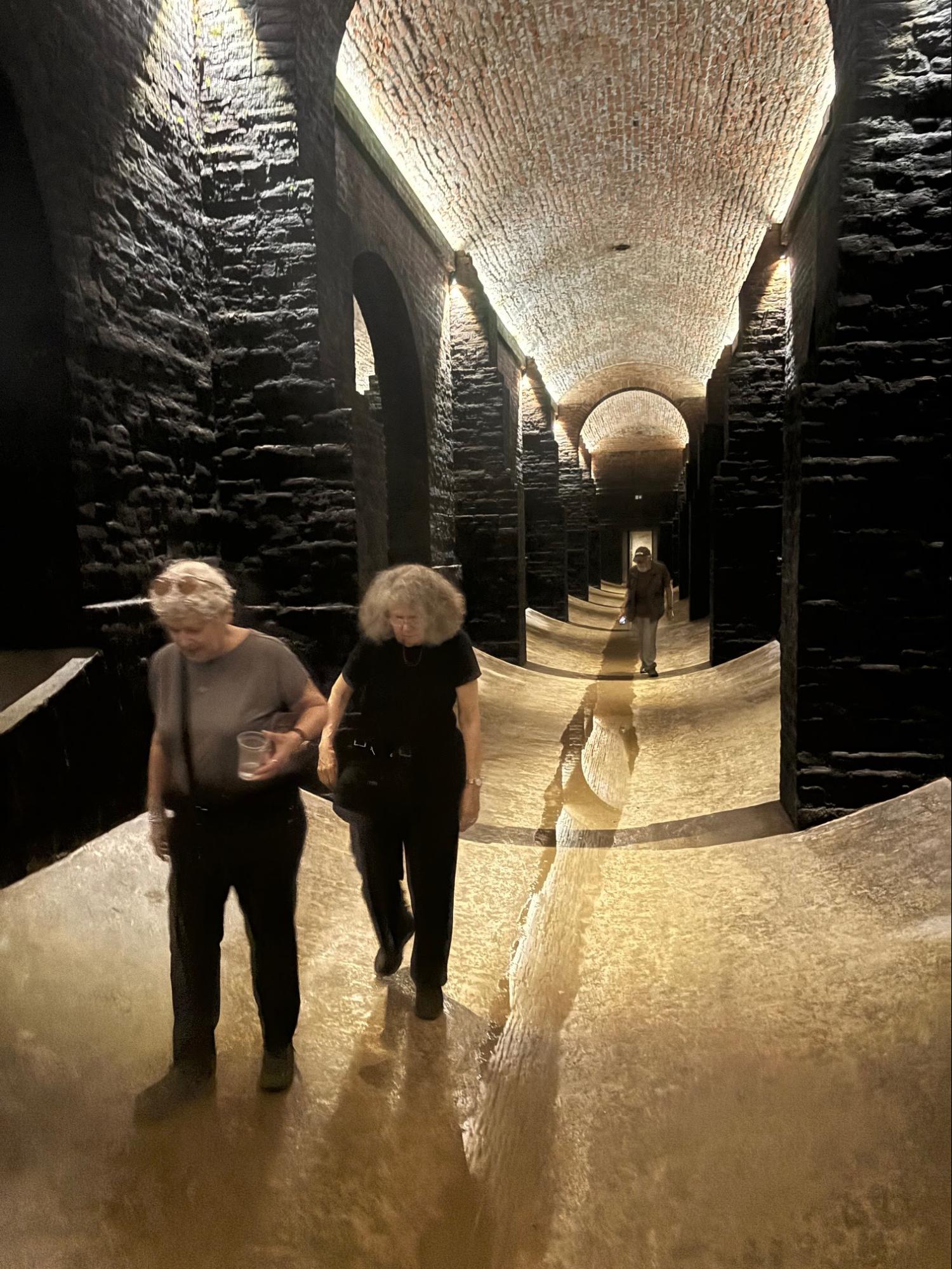 Two people walking through a dimly lit underground corridor with a curved floor covered in water, surrounded by dark brick walls and arched ceilings.