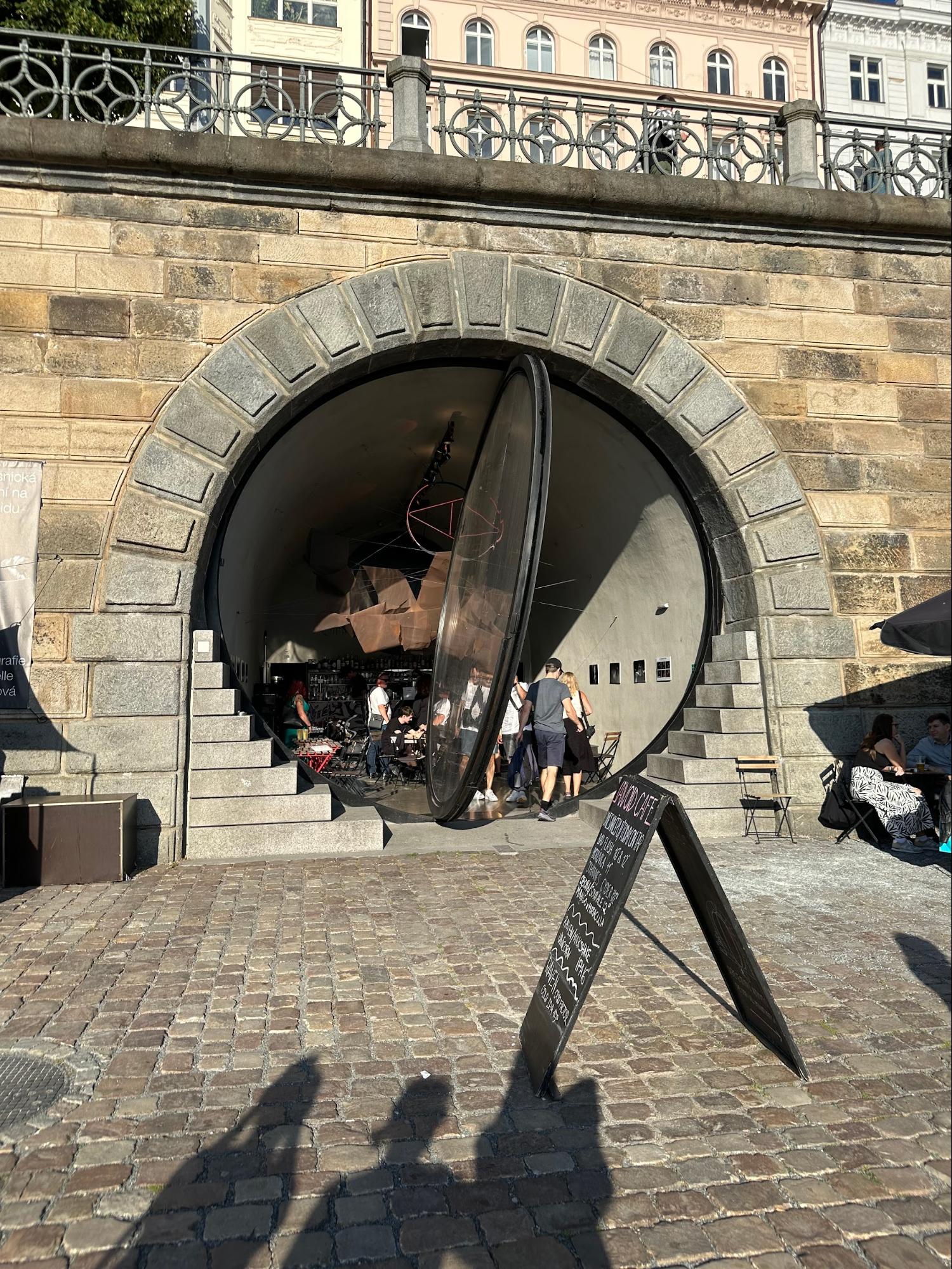 A large stone archway leads into a café with a glass door, and a chalkboard sign stands on the cobblestone street outside in the sunlight, with a few people walking in and out.