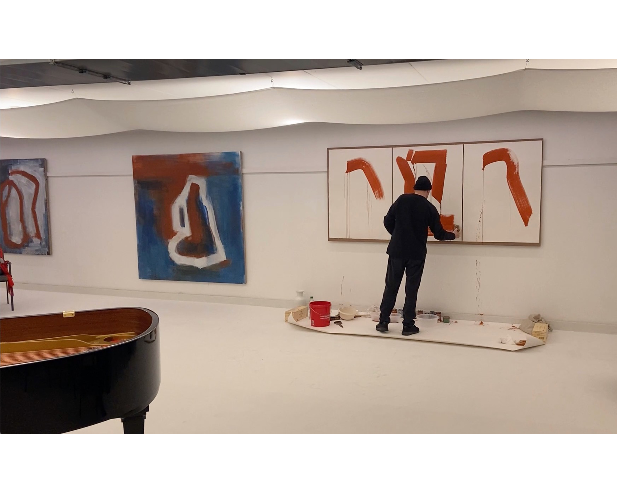 An artist dressed in black stands in a minimalist gallery space, painting bold red strokes on a triptych canvas mounted on the wall. Surrounding him are abstract paintings with deep blue, white, and red hues, while a grand piano in the foreground adds to the artistic ambiance of the room.