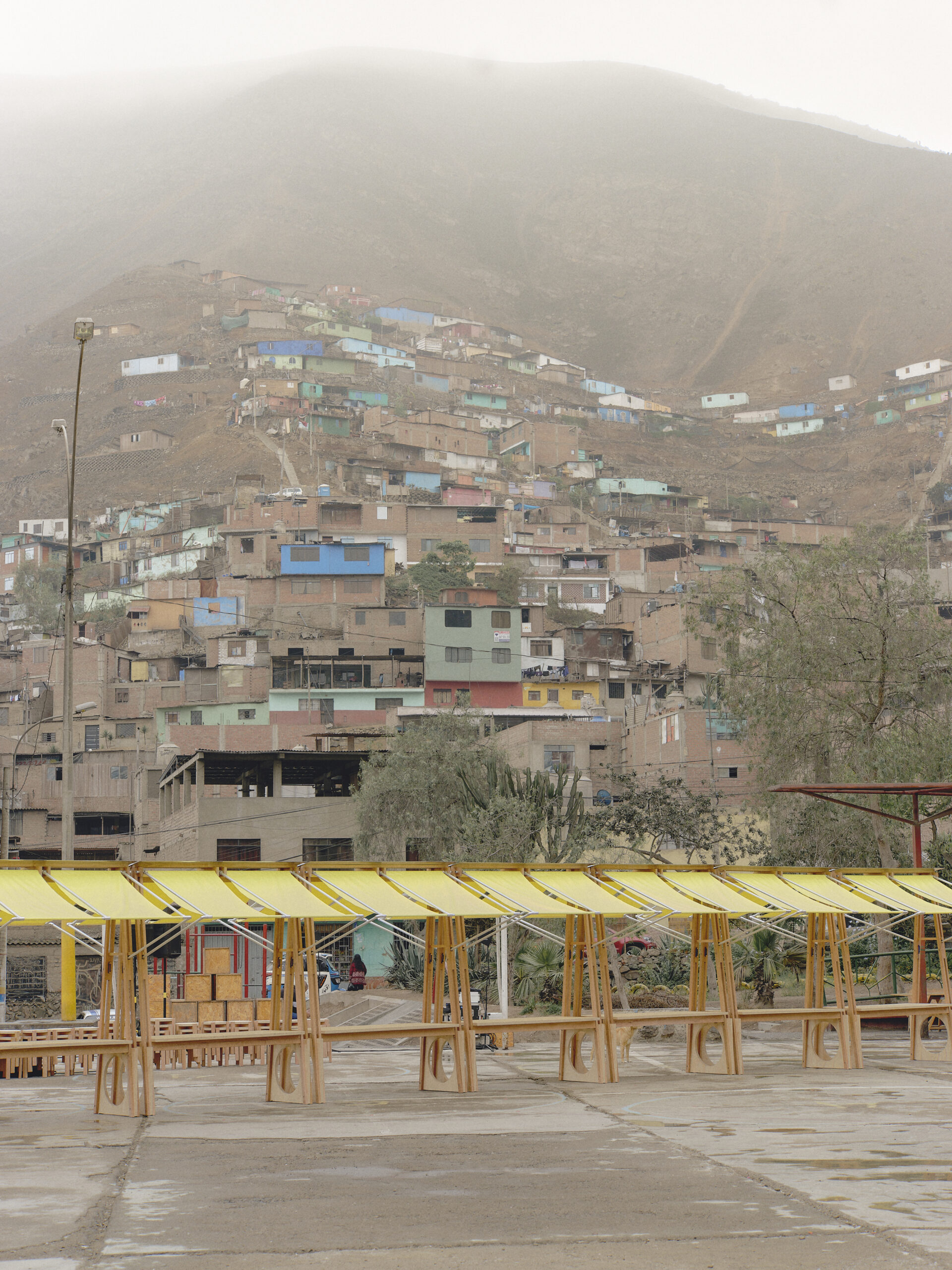 A hillside community with colorful houses is visible in the background, with mist covering the upper part of the mountain. In the foreground, there is a row of wooden structures with bright yellow canopies, possibly for a public space or market setup.