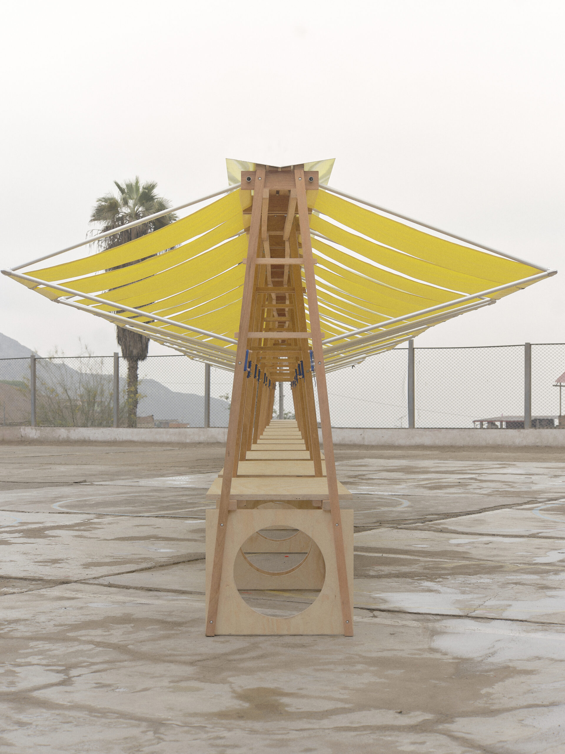 A close-up view of wooden structures with circular cutouts at the base, supporting bright yellow canopies stretched by white poles. In the background, a hillside community with stacked houses and palm trees are visible.