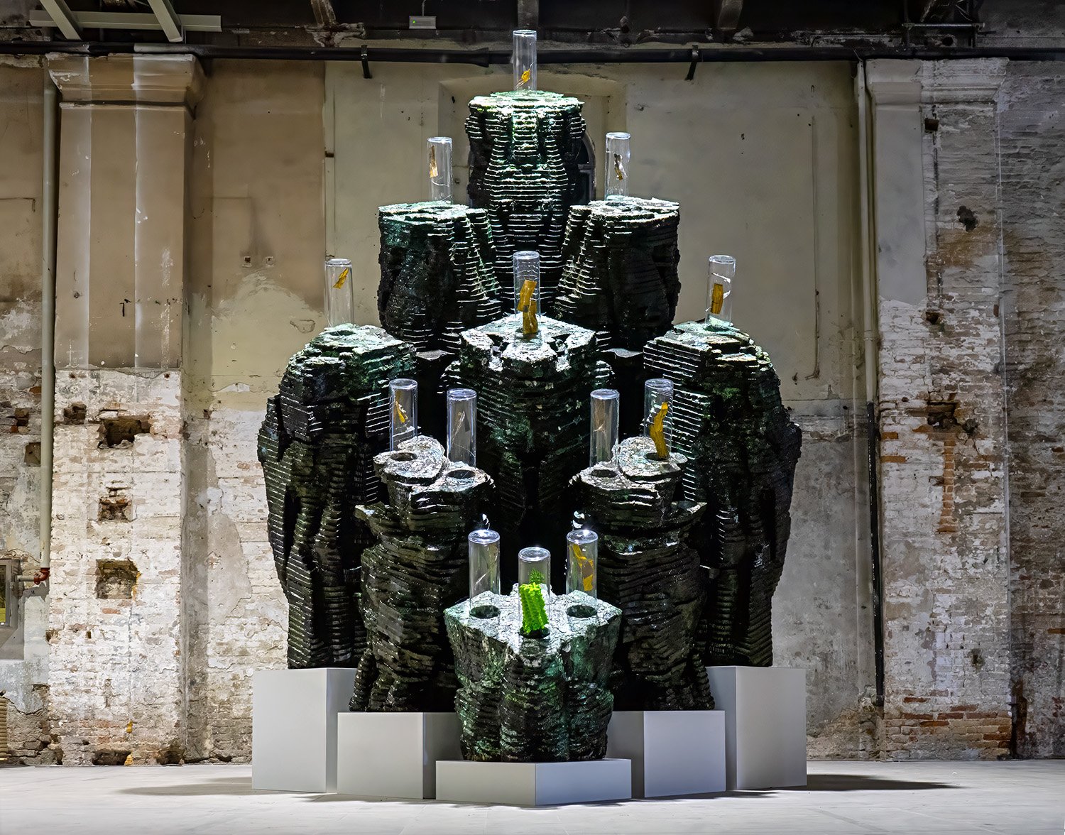 A sculptural installation of stacked, dark green, textured forms resembling abstract rock formations, each topped with a transparent cylinder containing preserved seaweed, displayed against a weathered brick and plaster wall.