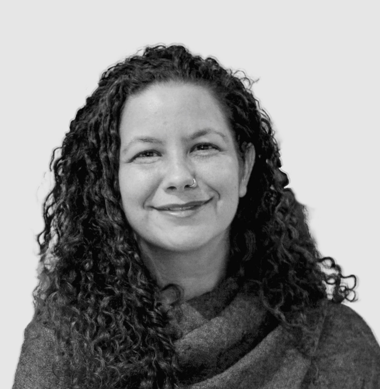 A black-and-white portrait of a person with long, curly hair, wearing a textured wrap-style top. They have a nose ring and a gentle smile, looking directly at the camera. The background is a smooth, light gray, creating a clean and professional setting for the image.