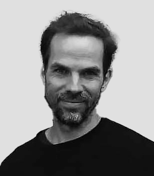 A black-and-white portrait of a person with short dark hair and facial hair, smiling slightly while looking at the camera. They wear a dark shirt and are positioned against a plain, light gray background, creating a minimal and focused composition.