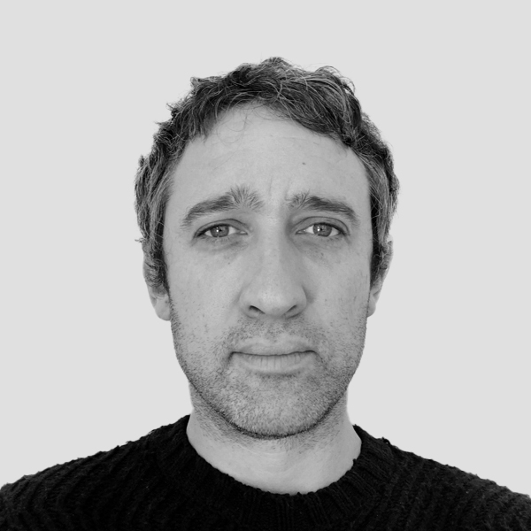 A black-and-white portrait of a person with short, curly hair and a serious expression, looking directly into the camera. They wear a textured, dark sweater and are set against a flat, light gray background, producing a straightforward and contemplative tone.