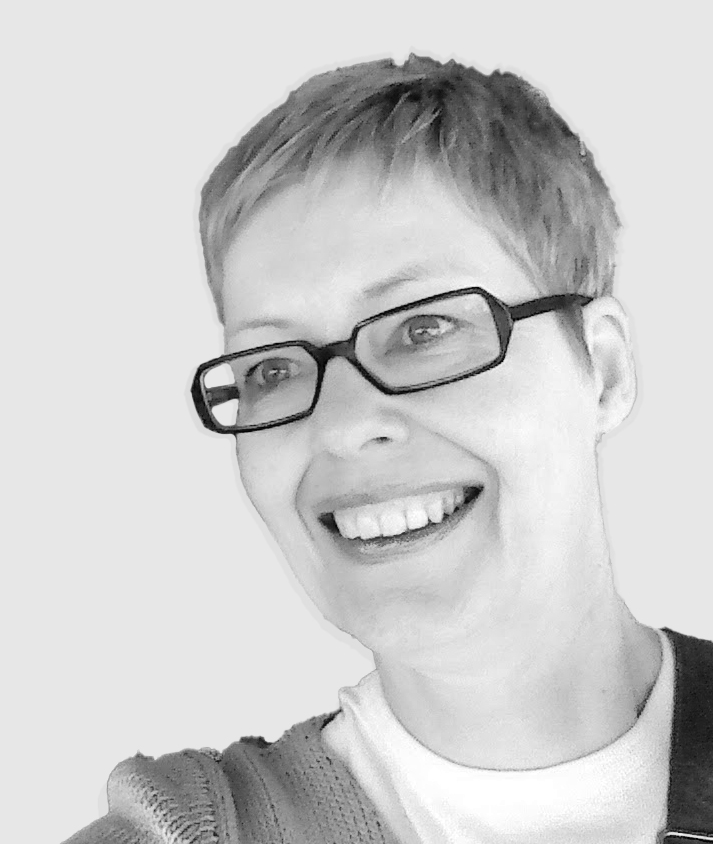 A black-and-white portrait of a person with short hair and rectangular glasses, smiling while looking slightly off camera. They wear a textured sweater and a white shirt underneath. The background is a solid light gray, giving the image a clean and professional appearance.