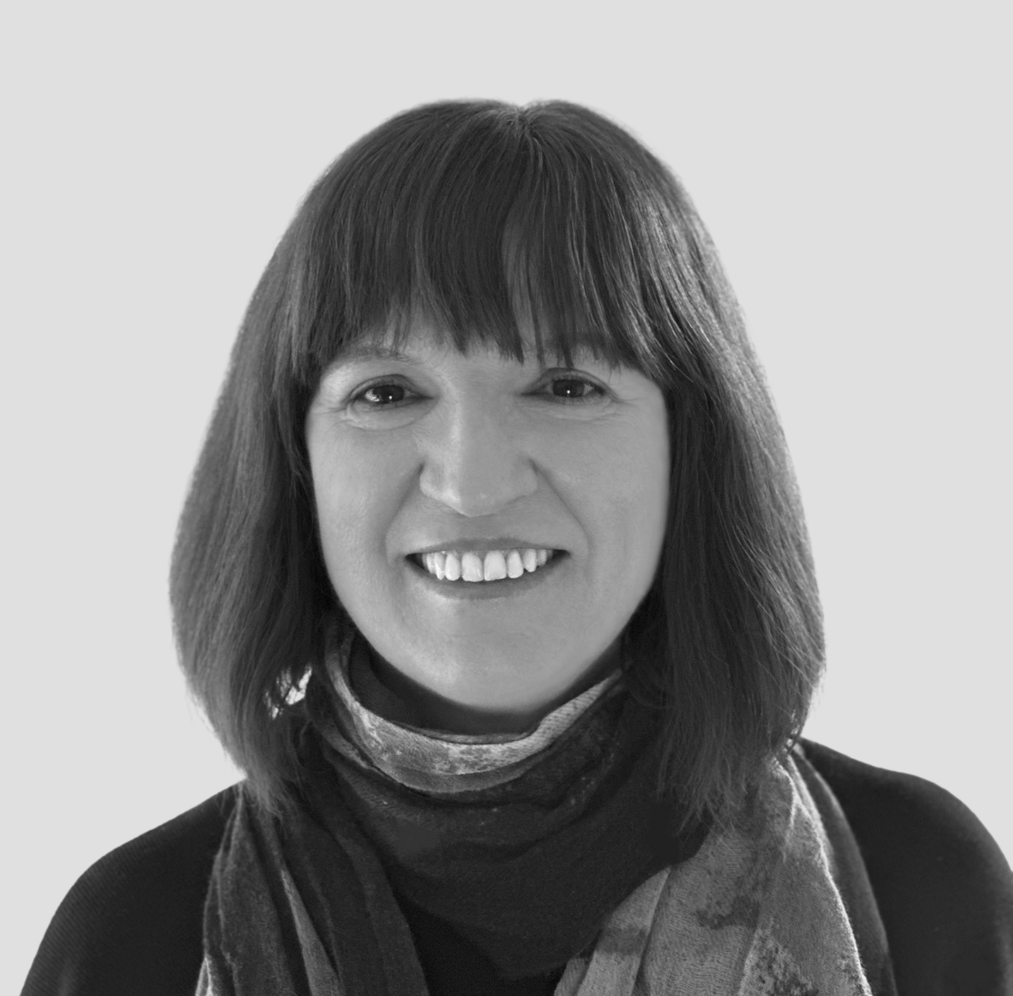 A black-and-white portrait of a person with shoulder-length hair and bangs, smiling at the camera. They are wearing a patterned scarf over a dark top. The background is light gray, presenting a formal and approachable profile.