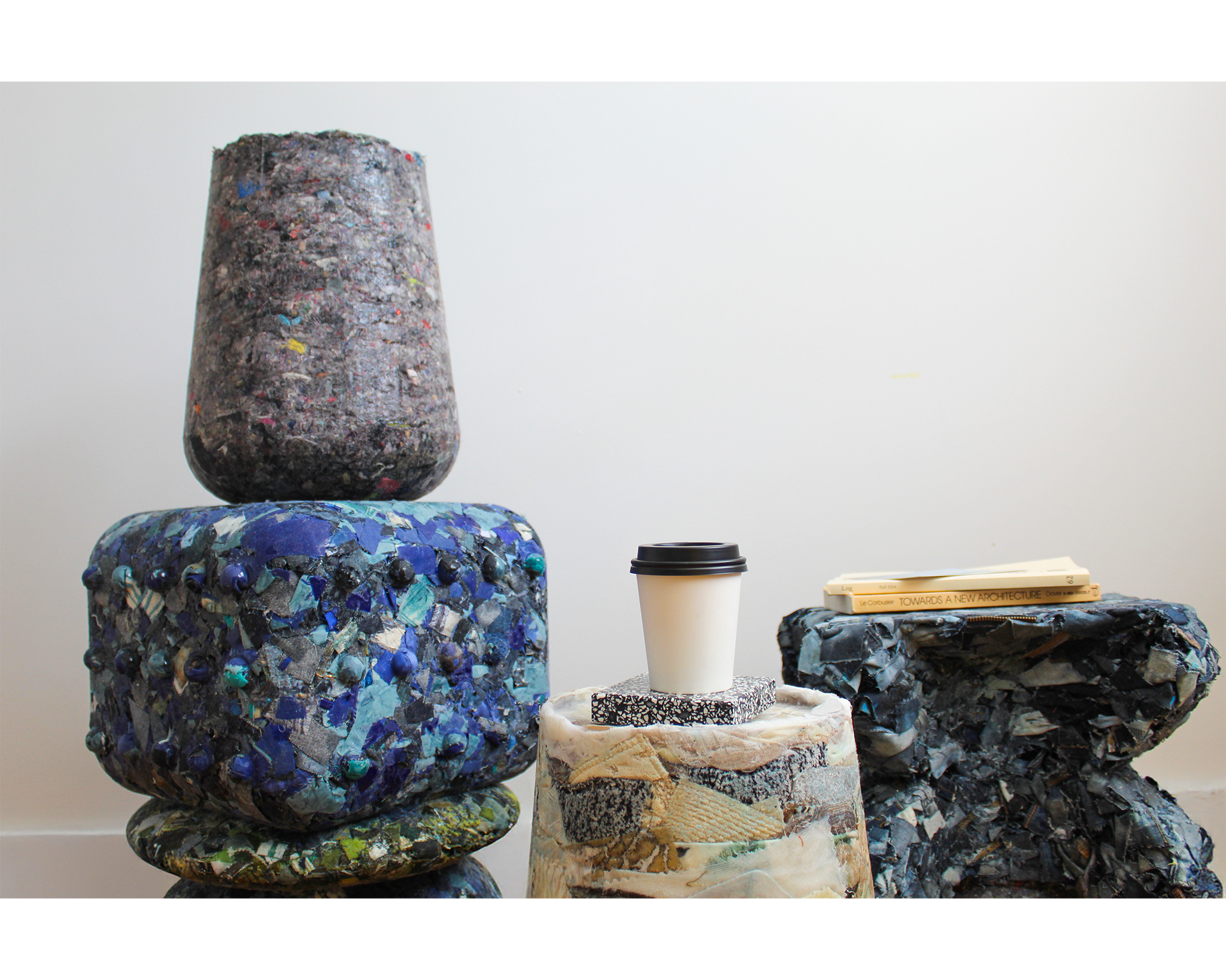 A group of sculptural furniture pieces made from recycled materials are arranged in a minimalist space, featuring textured surfaces in shades of blue, gray, and earth tones. On top of the pieces are a coffee cup, a patterned notebook, and two books including 