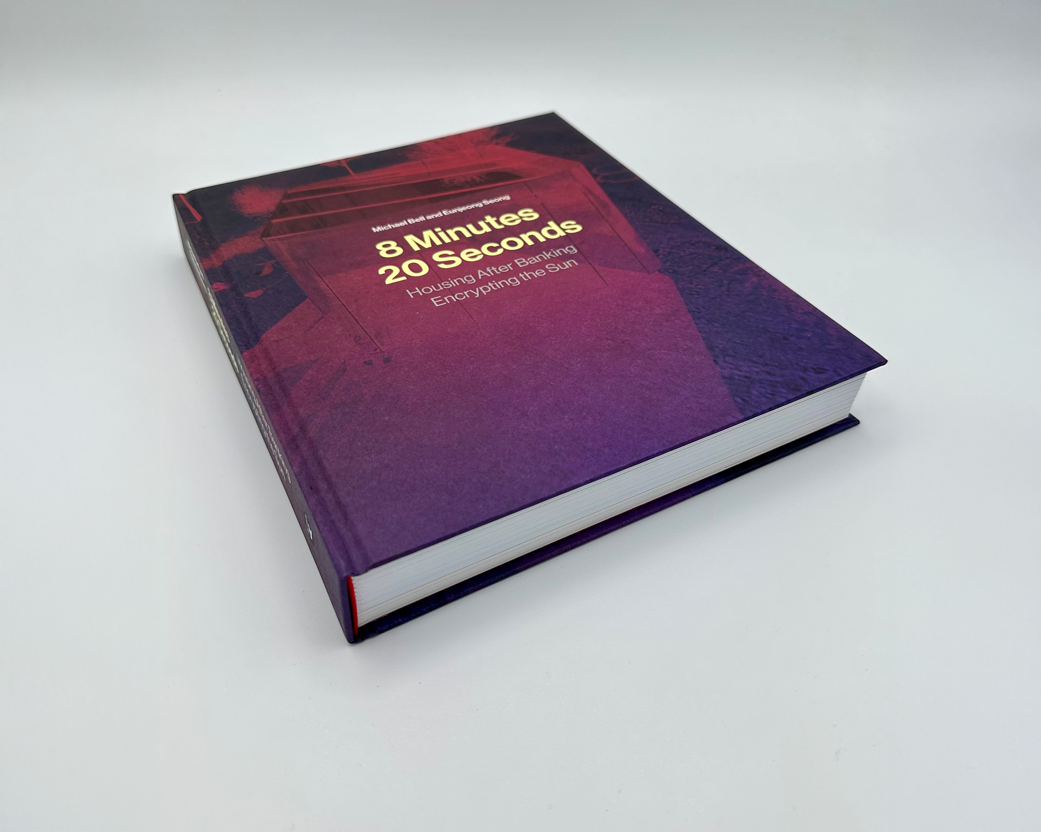 A hardcover book titled 8 Minutes 20 Seconds: Housing After Banking, Encrypting the Sun lies on a white surface. The cover features a purple and red abstract architectural image with gold and white text.