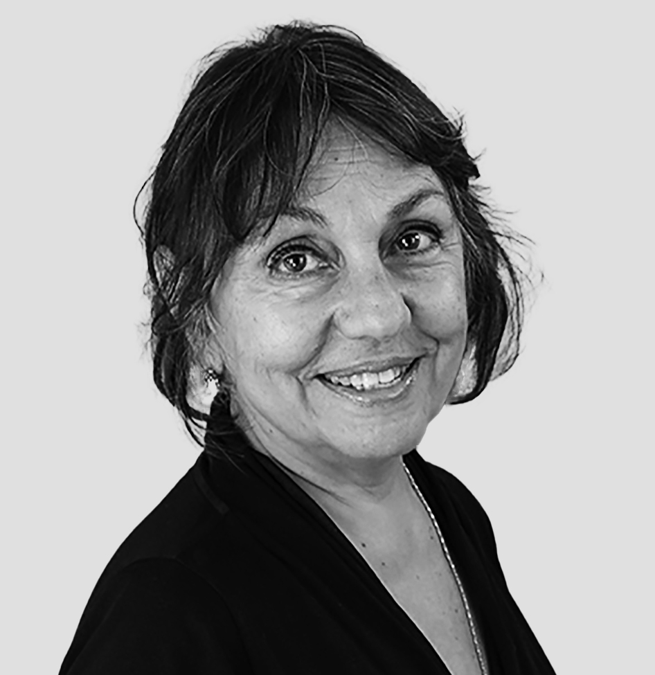 A black-and-white portrait of a person with dark hair loosely tied back, smiling warmly at the camera. They wear a dark v-neck top and small earrings. The background is plain and light gray, highlighting the subject clearly.