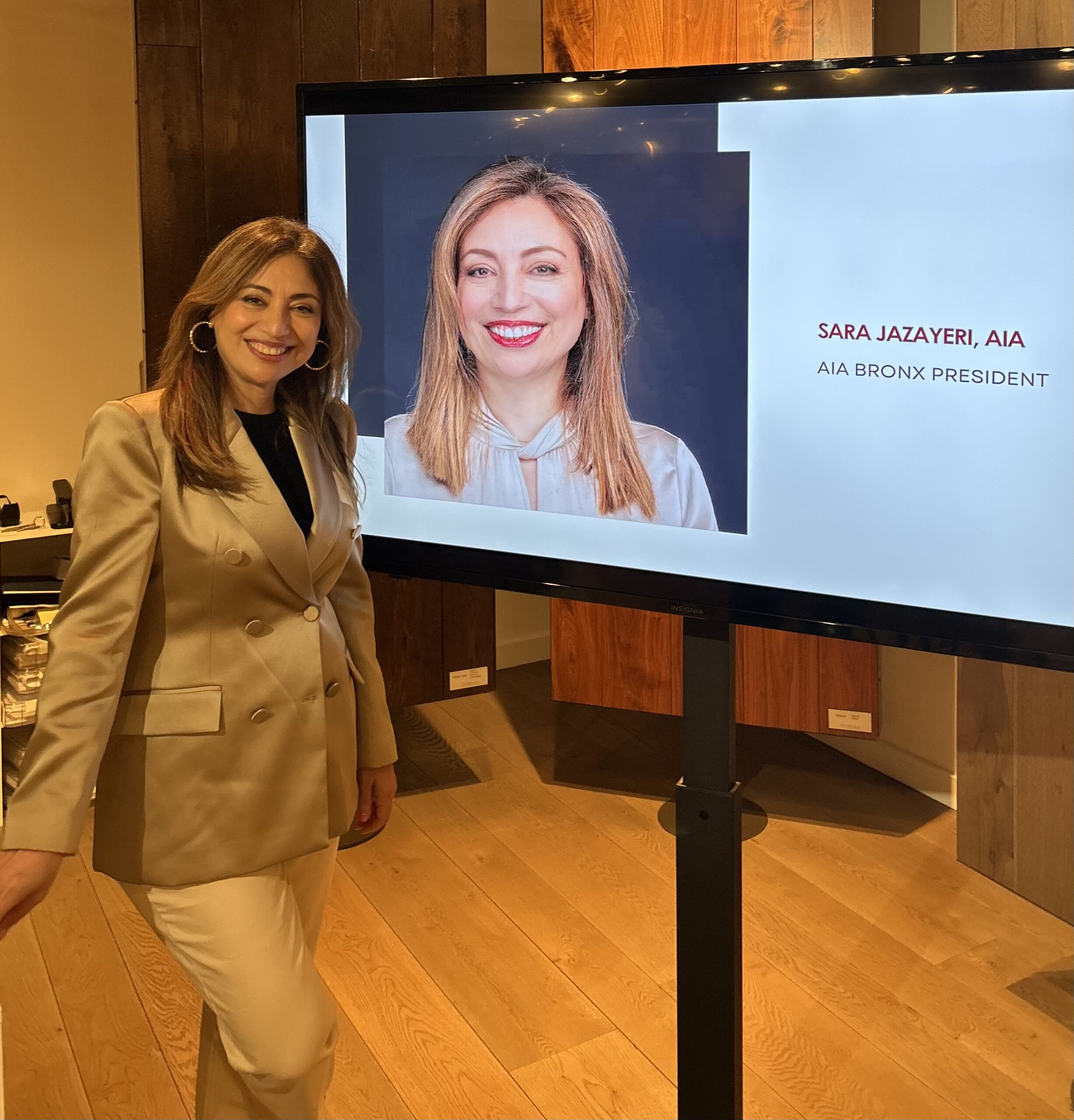 Sara Jazayeri stands smiling next to a screen displaying her portrait and title, "AIA Bronx President."