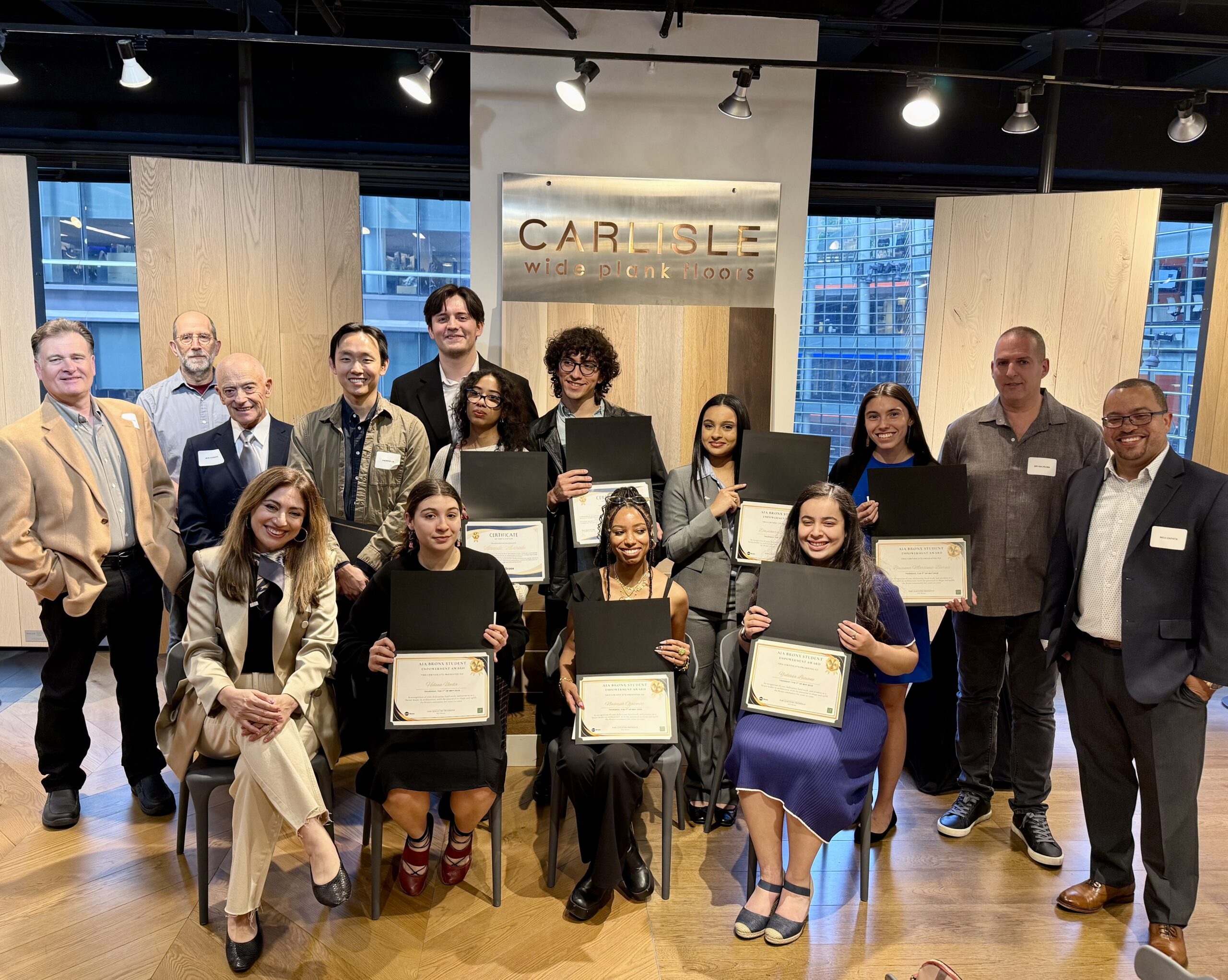 A group of scholarship recipients and presenters pose indoors with certificates in hand, smiling in front of a sign that reads “Carlisle Wide Plank Floors.”