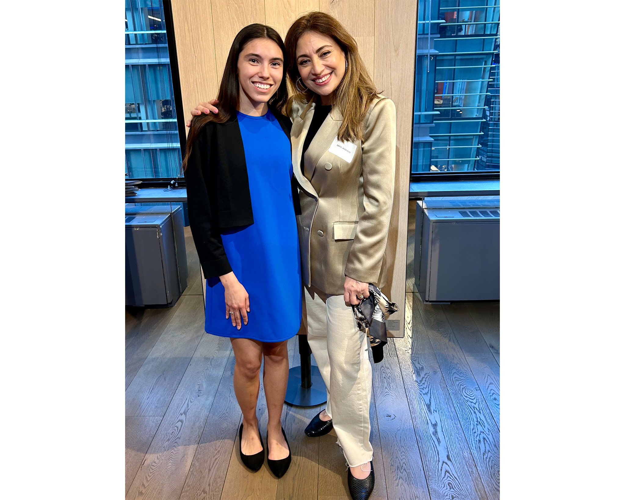 Two women stand smiling together indoors; one wears a bright blue dress with a black cardigan, and the other wears a gold blazer with white pants, holding a small handbag.