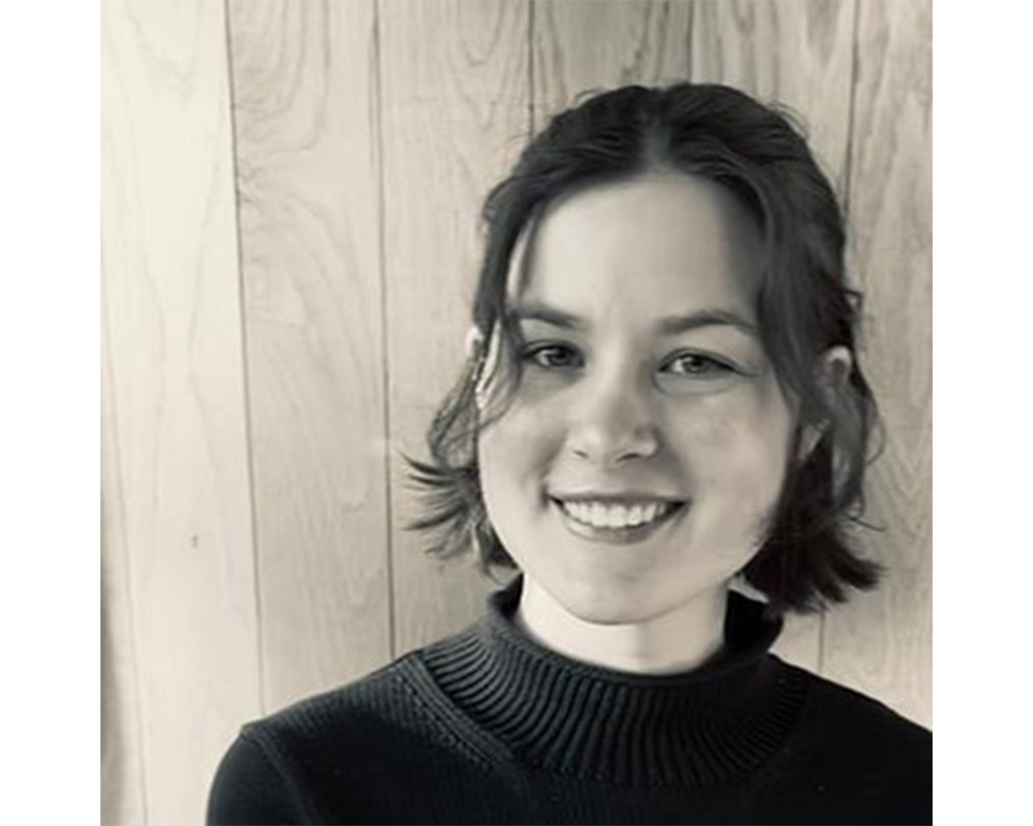 Black and white portrait of a woman in a turtleneck posed in front of a wood panel wall.