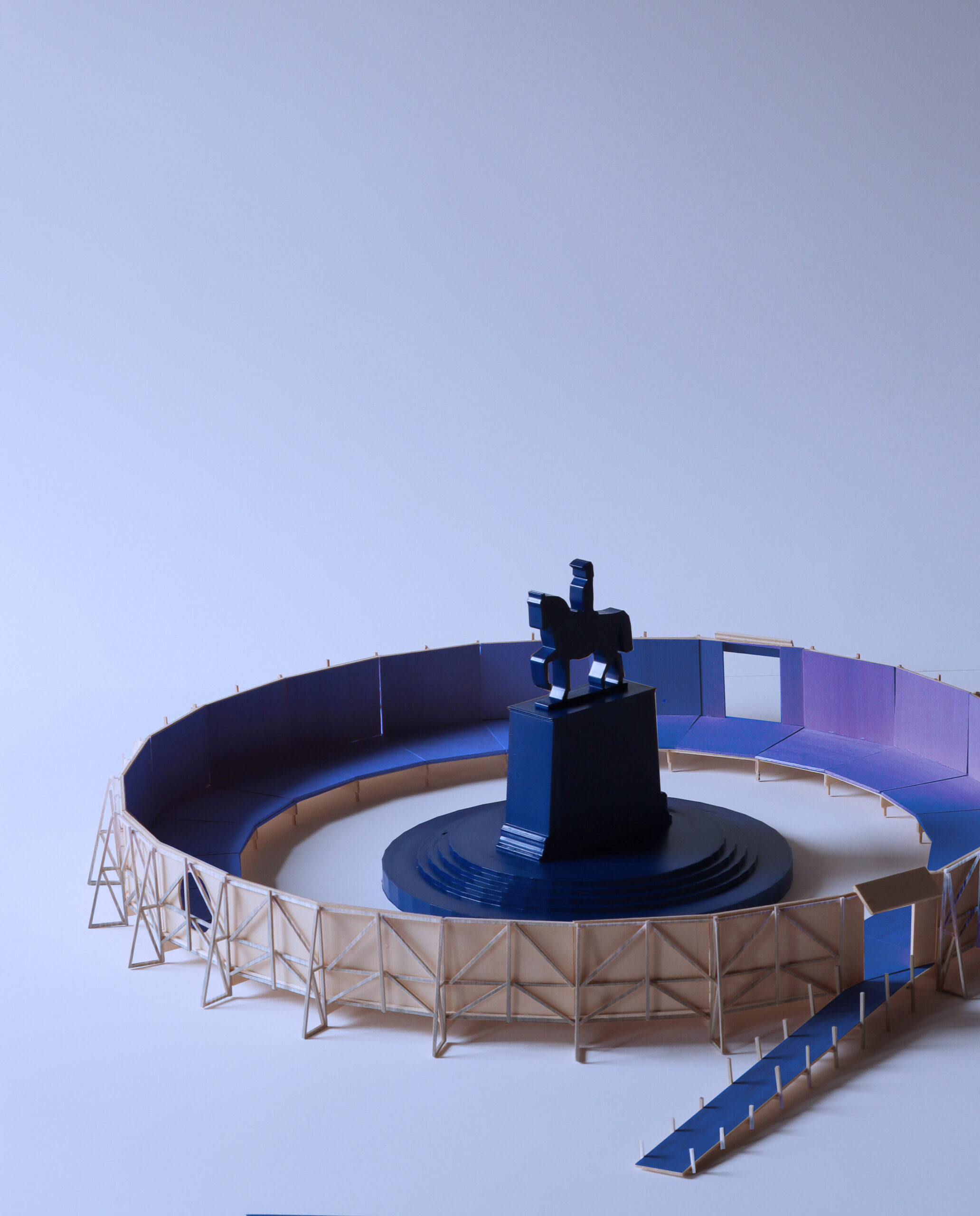 A scale model of a circular installation features a blue equestrian statue at the center, surrounded by a raised walkway and ramp enclosed by translucent structural supports.