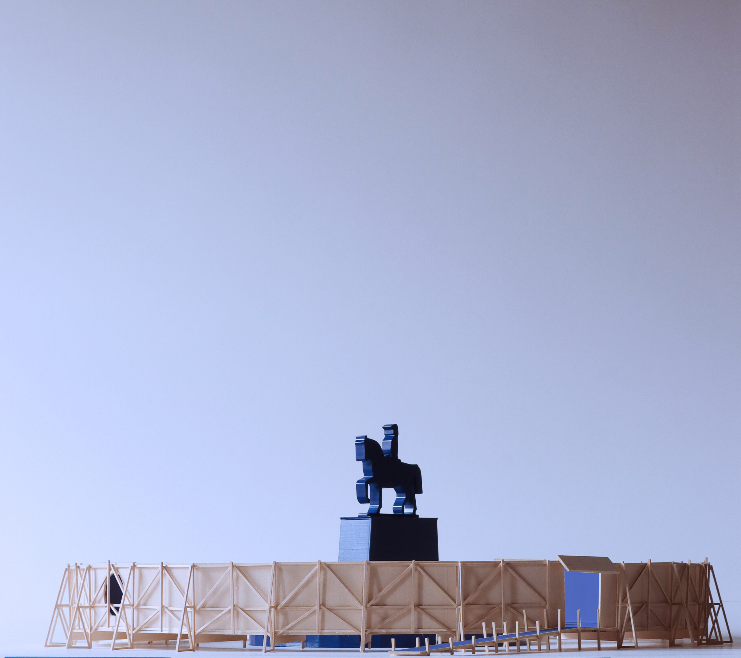 A scale model of a circular installation features a blue equestrian statue at the center, surrounded by a raised walkway and ramp enclosed by translucent structural supports.