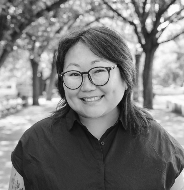 A woman with glasses smiles while standing on a tree-lined pathway in an outdoor setting.