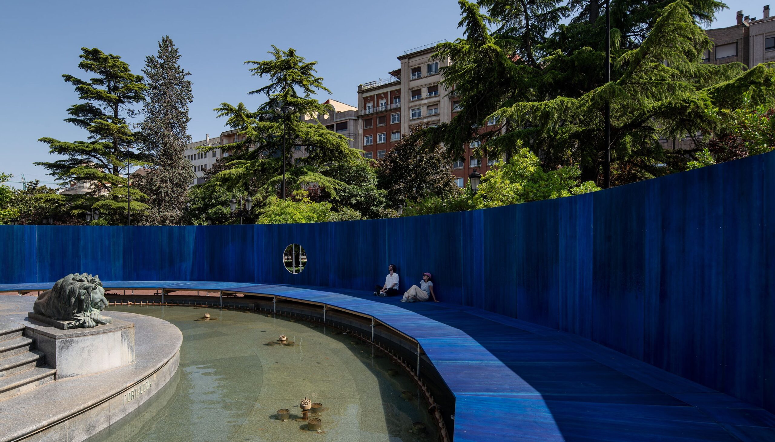 Two people sit along a curved, elevated blue walkway encircling a shallow fountain and lion sculpture, framed by trees and urban buildings in the background.