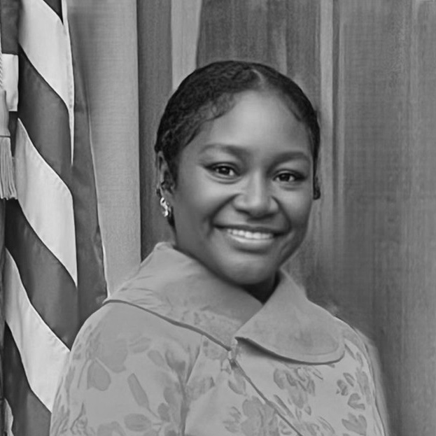 A woman smiles while standing in front of an American flag and wood-paneled background, wearing a patterned coat with a wide collar.