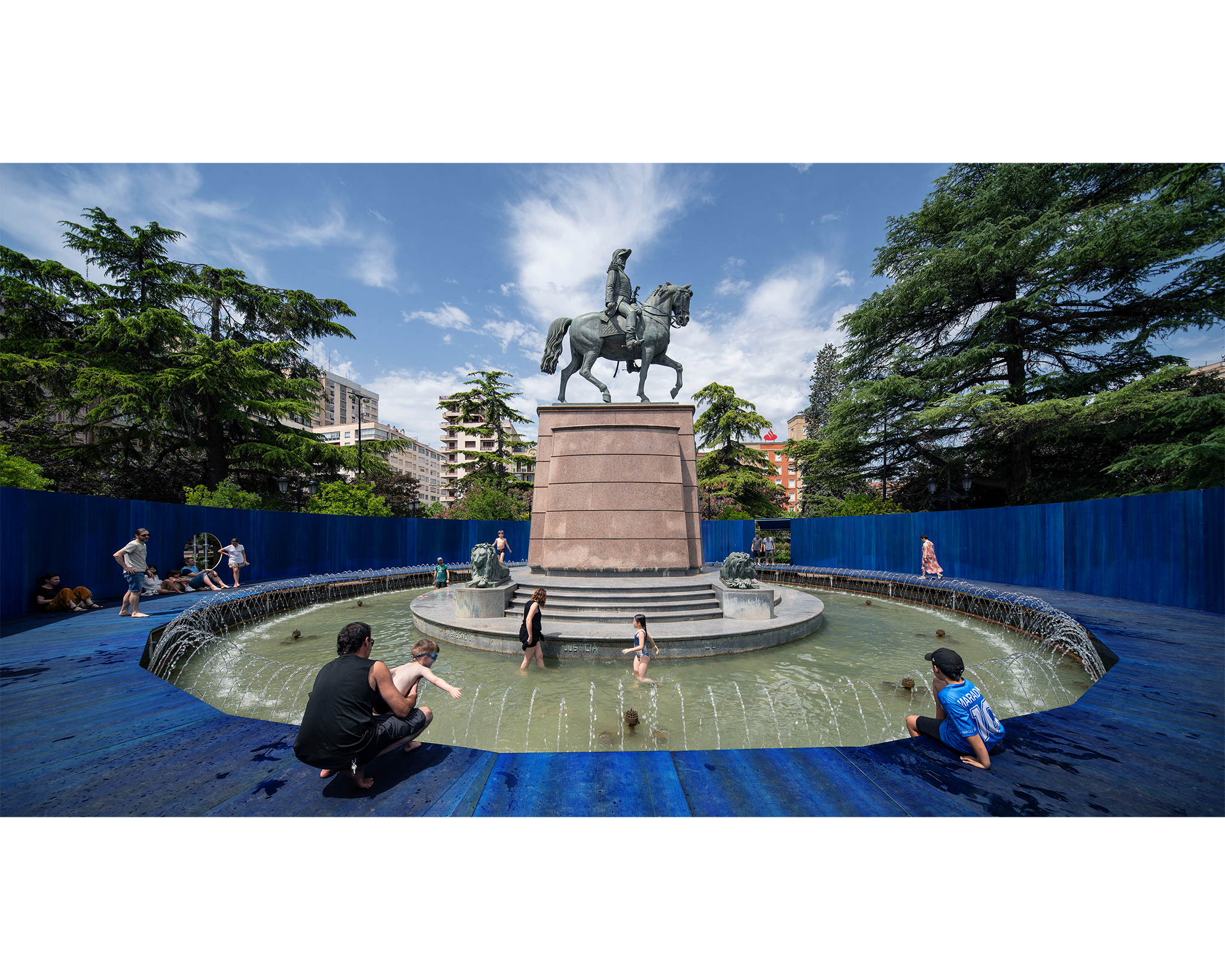 Visitors wade and relax in a circular fountain surrounding an equestrian statue, set within a vivid blue installation in a tree-lined urban plaza.