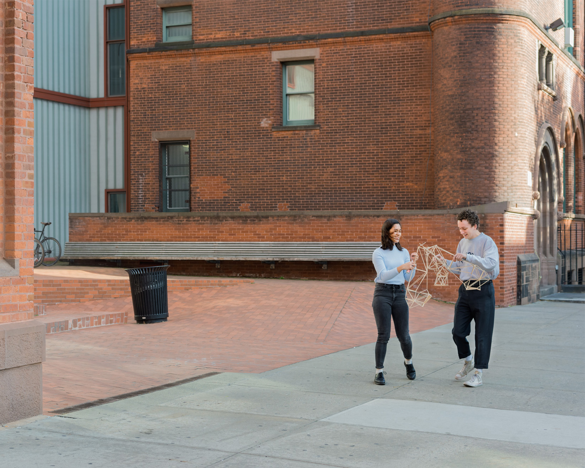 Students holding an architectural model walk in front of the Pratt Institute School of Architecture, Higgins Hall building.