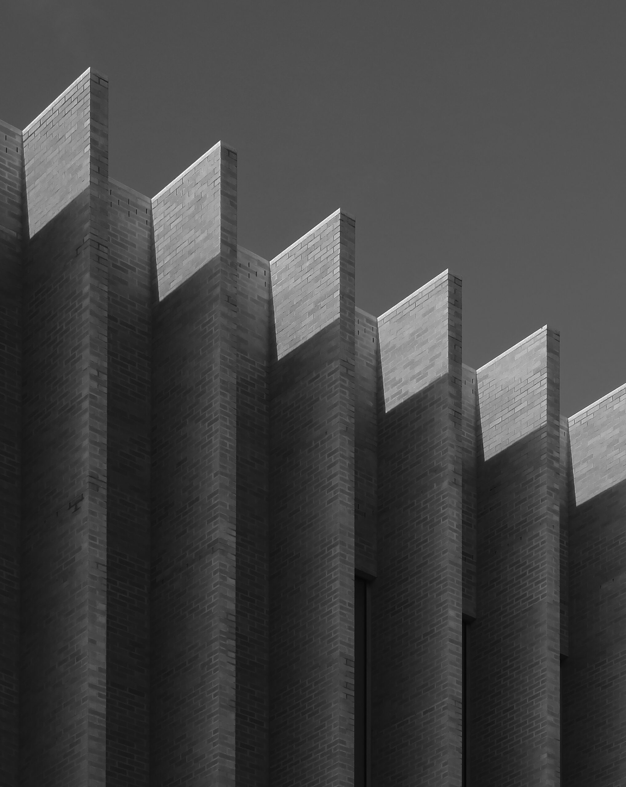 The image features a close-up view of a building's brick facade in black and white. The structure exhibits a series of angular protrusions and shadows, creating a striking geometric pattern that emphasizes the texture of the bricks and the interplay of light and dark. The sky in the background is clear and contrasts with the architectural elements.