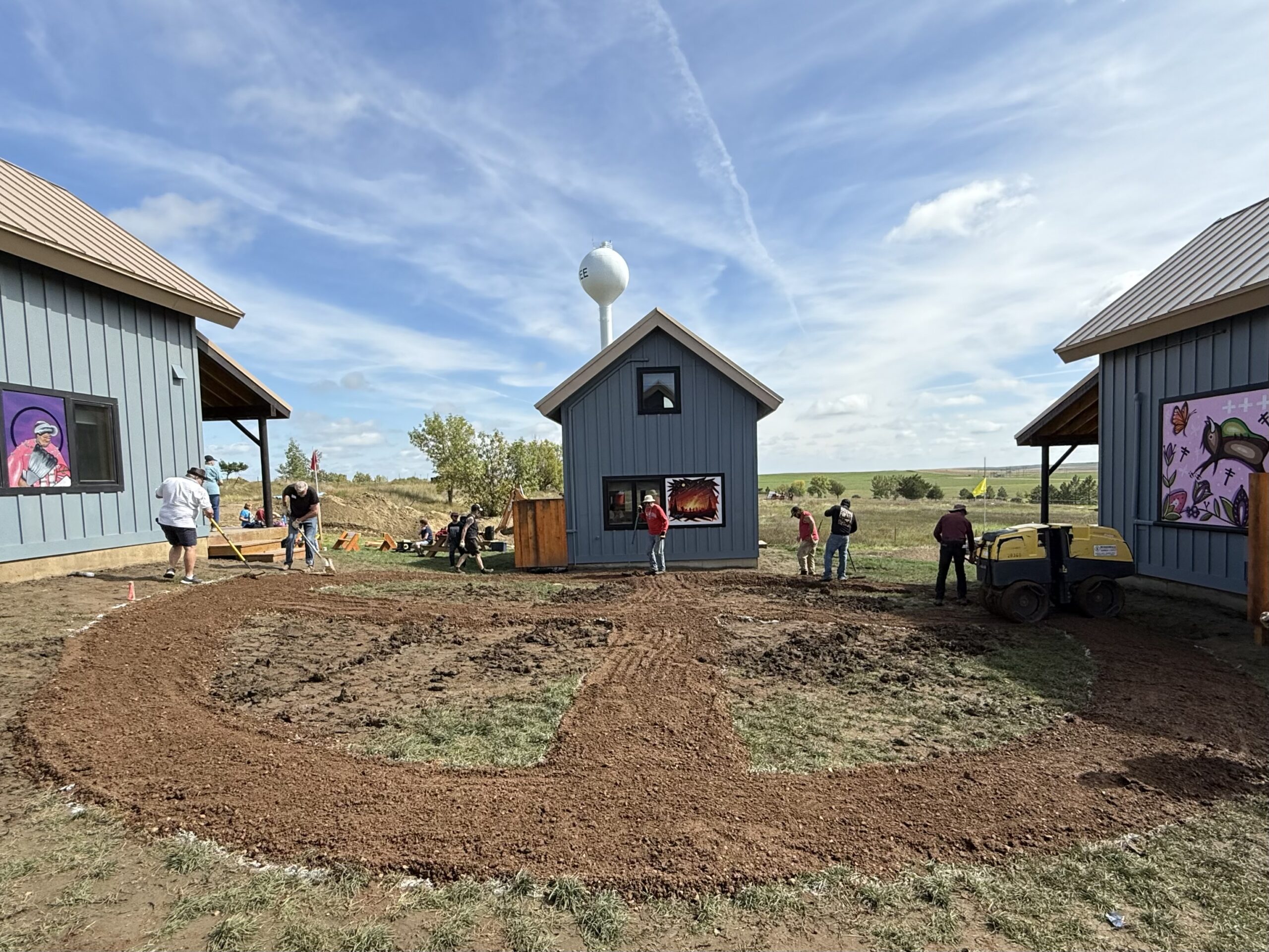 A landscaped area is being worked on between two blue buildings with metal roofs. Several people are engaged in various activities, such as digging and leveling soil. A water tower is visible in the background, along with artwork on the buildings and greenery in the surrounding area. The sky is partly cloudy, indicating a pleasant day.