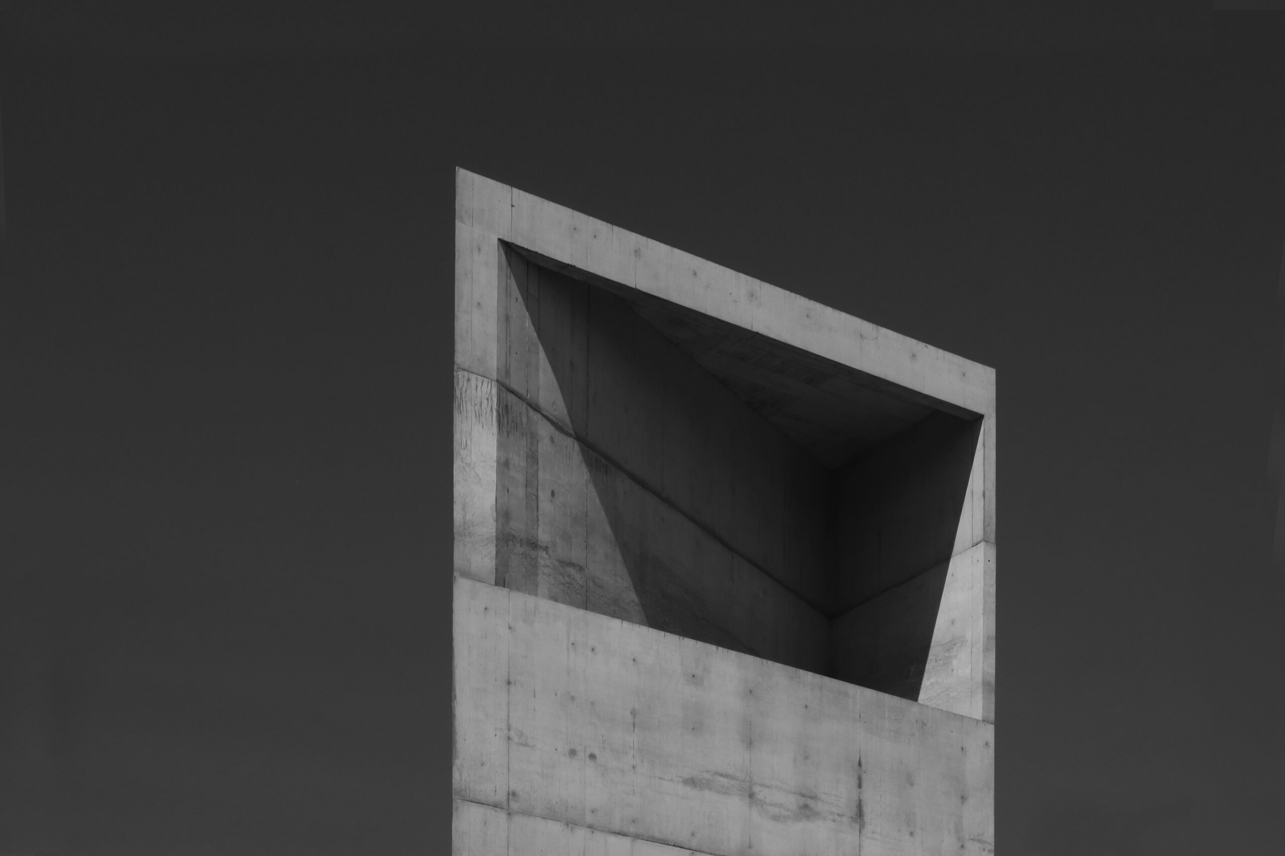 A tall, angular concrete structure rises against a dark sky. The design features sharp edges and geometric shapes, with a prominent overhang creating a shadowed area. The surface is textured and has a monochromatic appearance, emphasizing the interplay of light and shadow on the concrete.