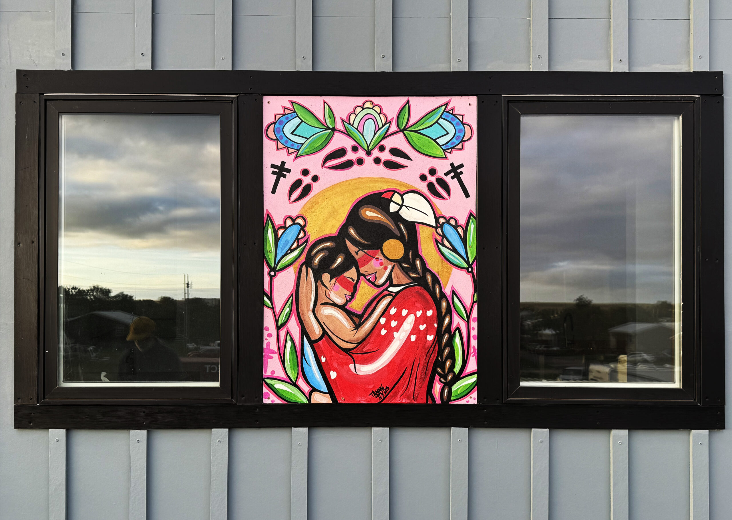 A colorful mural depicts two figures embracing, surrounded by vibrant flowers and leaves. The background features a pink hue with a sun motif, adding warmth to the scene. The figures have bold outlines, with one wearing a red dress and the other appearing smaller, creating an intimate moment. Reflections of outdoor scenery can be seen in the glass windows framing the mural, enhancing the visual interest of the artwork against a gray wall.