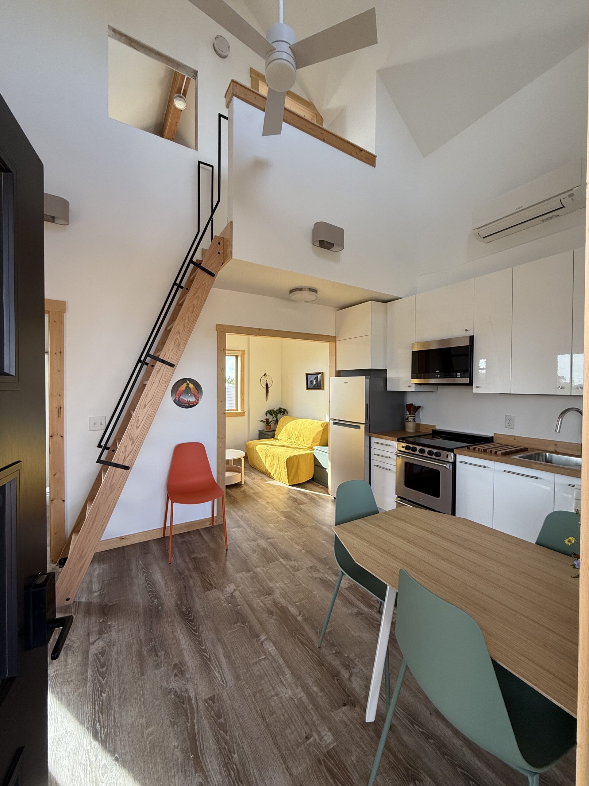 A modern interior of a small apartment featuring a staircase leading to an upper level. The main area includes a dining table with green and white chairs, a bright orange chair, and a living space with a yellow sofa. The kitchen appliances are sleek and minimalistic, with a stovetop, oven, and refrigerator visible. Large windows provide natural light, and wooden accents enhance the design. The flooring is a warm, light brown wood.