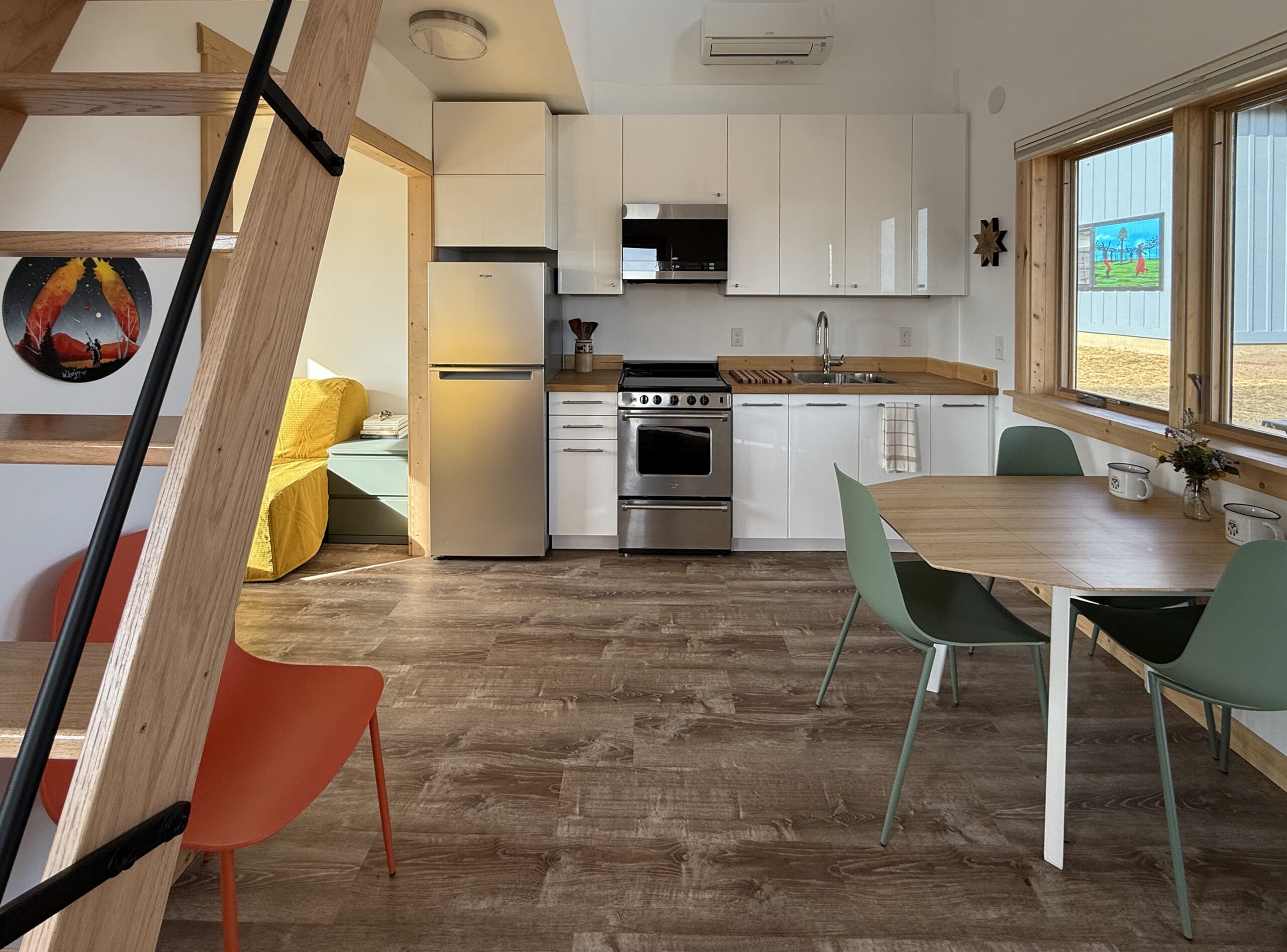 A modern compact kitchen and dining area featuring a stainless steel refrigerator, stove, and sink with wooden countertops. There are light-colored cabinets above, and a dining table with green chairs and white mugs. An orange chair is visible in the foreground. A cozy yellow sofa is seen through an open doorway. Natural light streams through large windows, illuminating the wooden flooring and minimalistic decor.