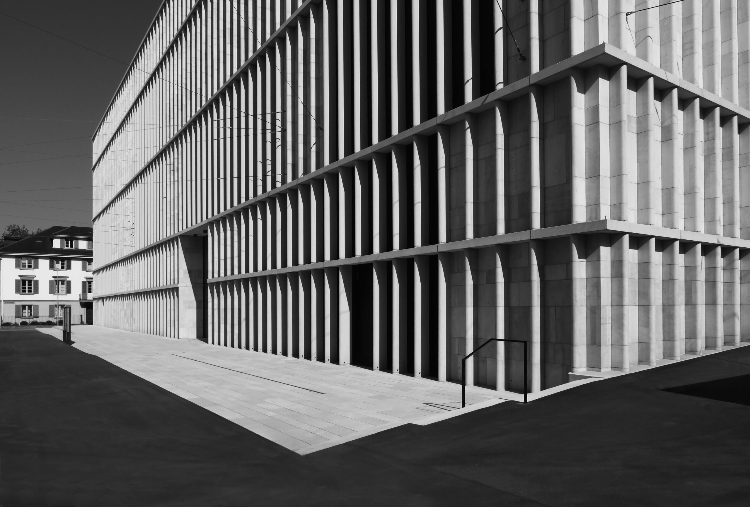 A modern building façade featuring a series of vertical columns and slits, creating a rhythmic pattern with varying light and shadow. The structure is primarily in shades of gray, and the foreground has a smooth paved surface leading to a simple staircase with a black handrail. In the background, a smaller traditional building with windows can be spotted, contrasting the sleekness of the modern architecture.