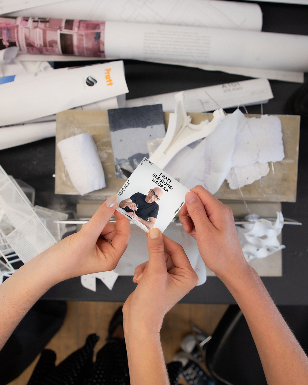 Two people hold a small printed card featuring the “Pratt Sessions: NADAAA” promotional image of a man writing in a notebook. The background shows an architecture studio workspace with models, drawings, and Pratt-branded paper rolls.