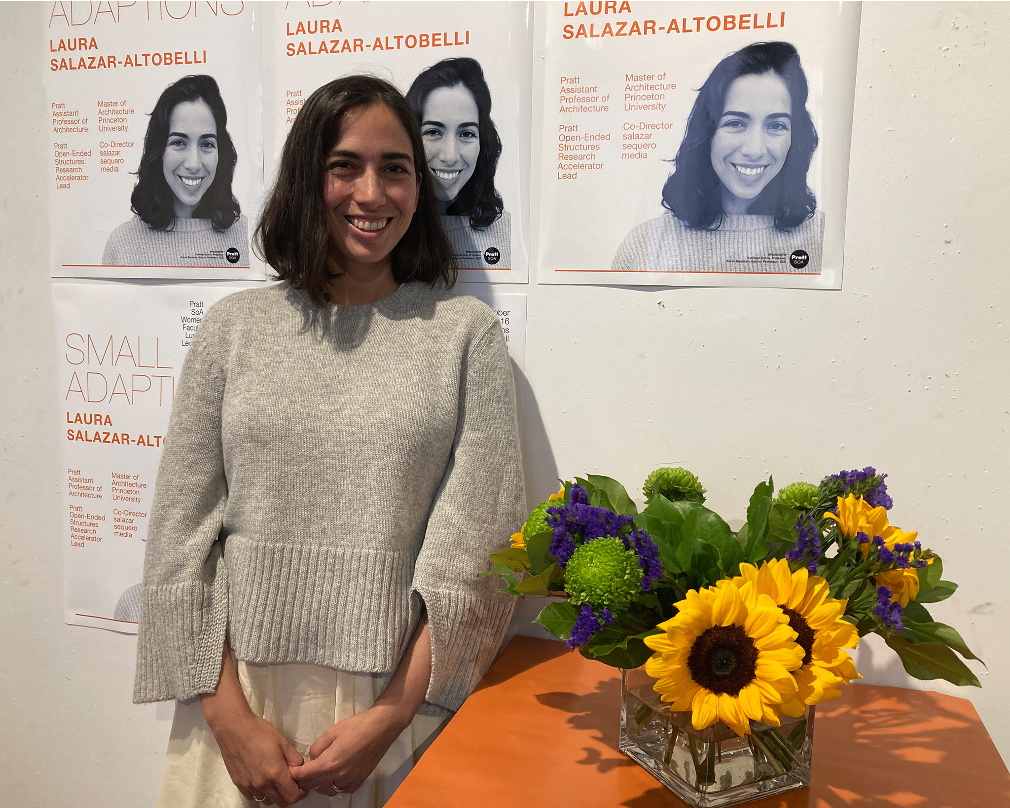 Laura Salazar-Altobelli stands next to a vase of flowers, in front of the event poster for the SoA Women Faculty Lunch and Lecture.