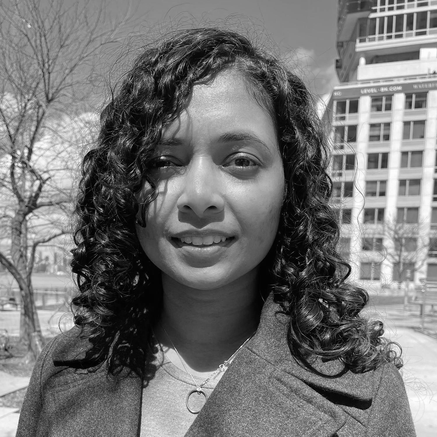 A woman with curly hair is smiling at the camera, wearing a dark coat over a lighter top. The background features bare trees and a modern building. The image is in black and white, highlighting her facial expression and the textures of her outfit.