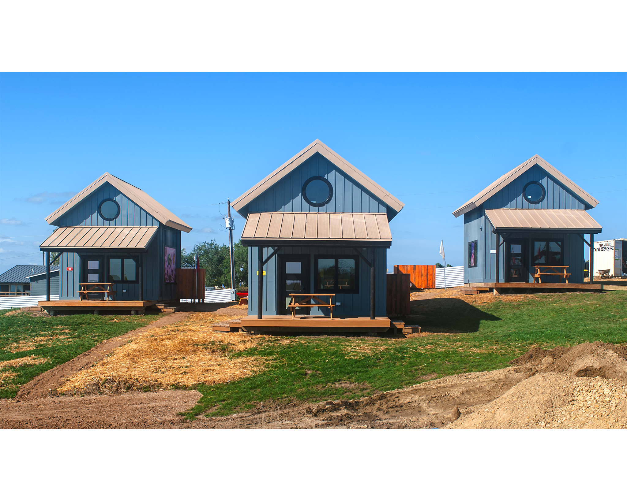 Three modern, blue-painted tiny houses with triangular roofs are situated on a grassy area. Each house has a porch with seating. The sky is clear and sunny, with a few scattered clouds, while the ground features some straw and earth patches.