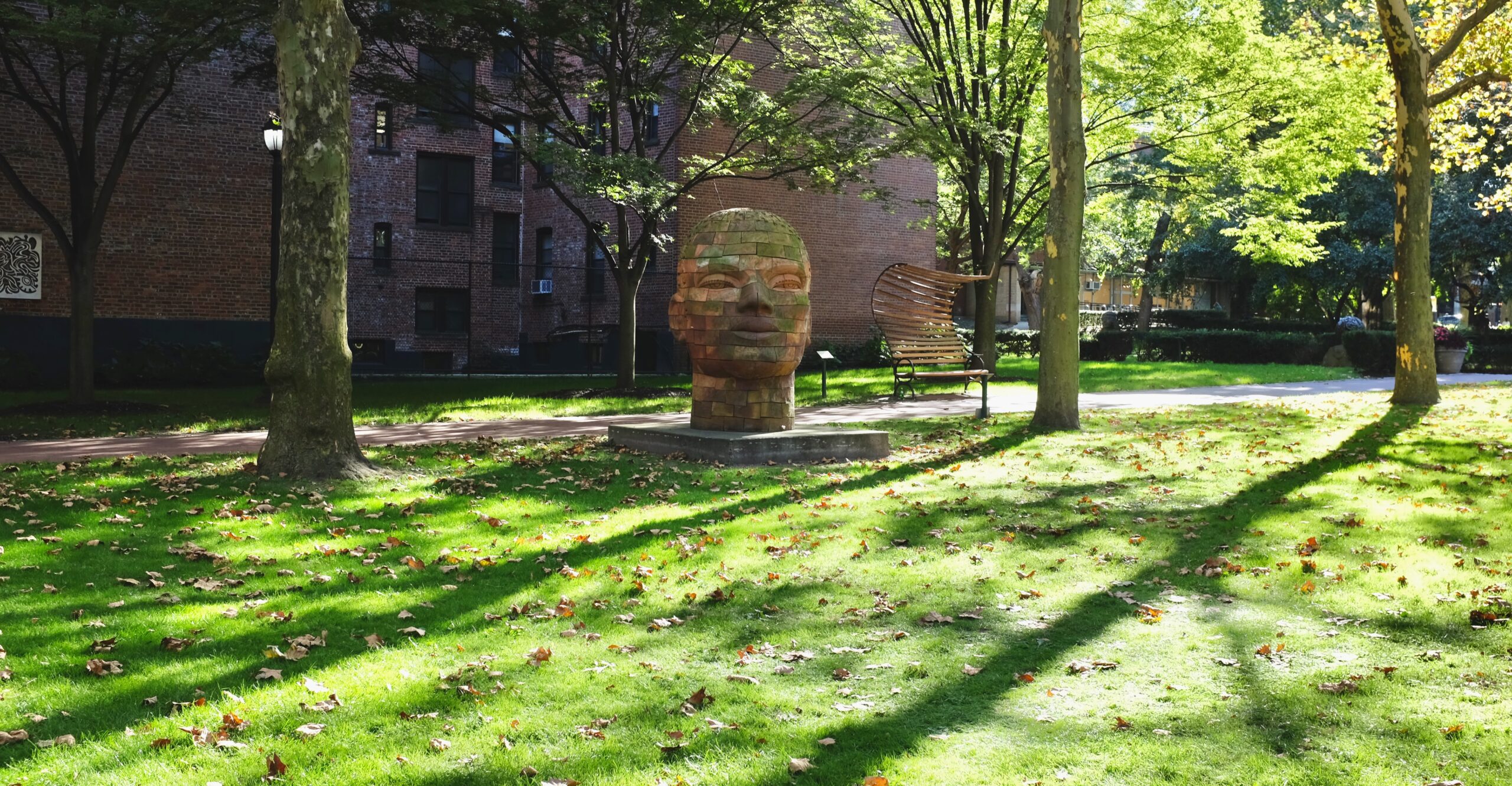 A large stone sculpture of a face is centered in a grassy park area, surrounded by trees and fallen leaves. Sunlight filters through the leaves, creating dappled shadows on the ground. In the background, there is a brick building and a wooden bench with a unique, curved design.