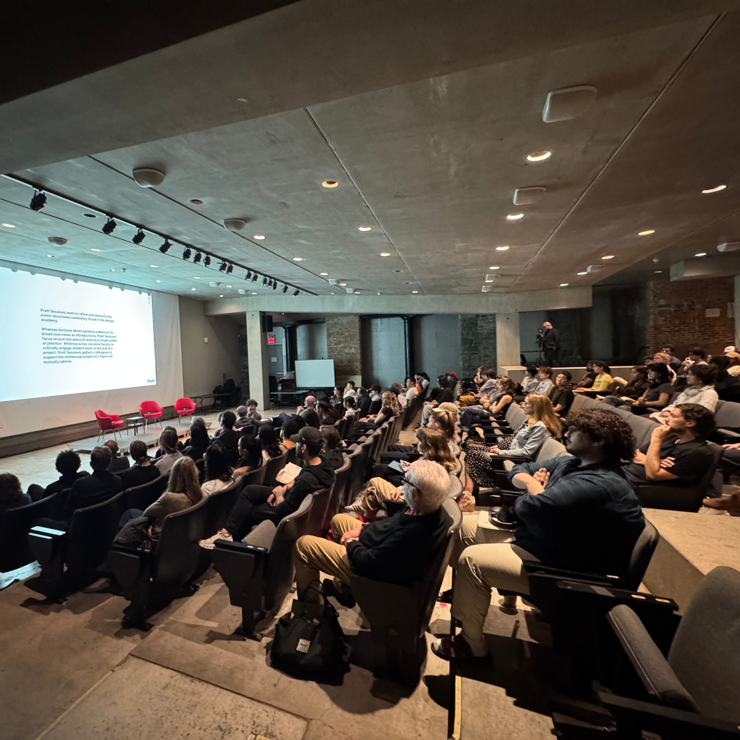 Audience members seated in an auditorium at Pratt Institute, facing a large projection screen displaying text about the Pratt Sessions lecture series. The room is well-lit, with rows of attendees focused on the presentation.