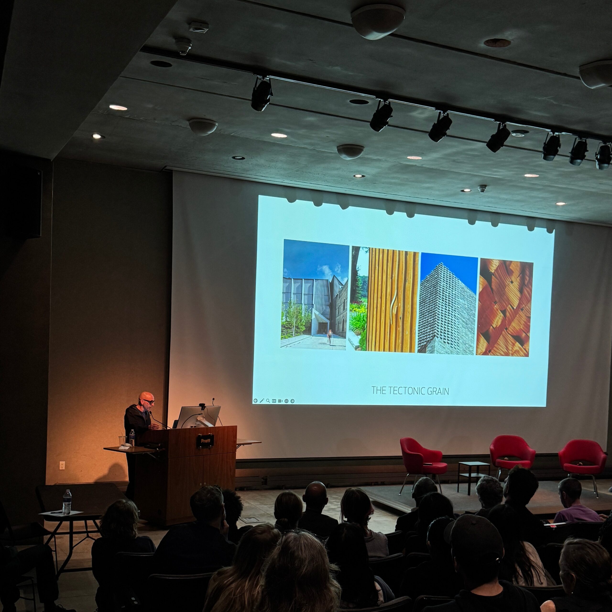 A speaker stands at the Pratt Institute podium during a lecture, presenting a slide titled “The Tectonic Grain” that displays four architectural images. The audience sits in dim lighting as they view the projected visuals.