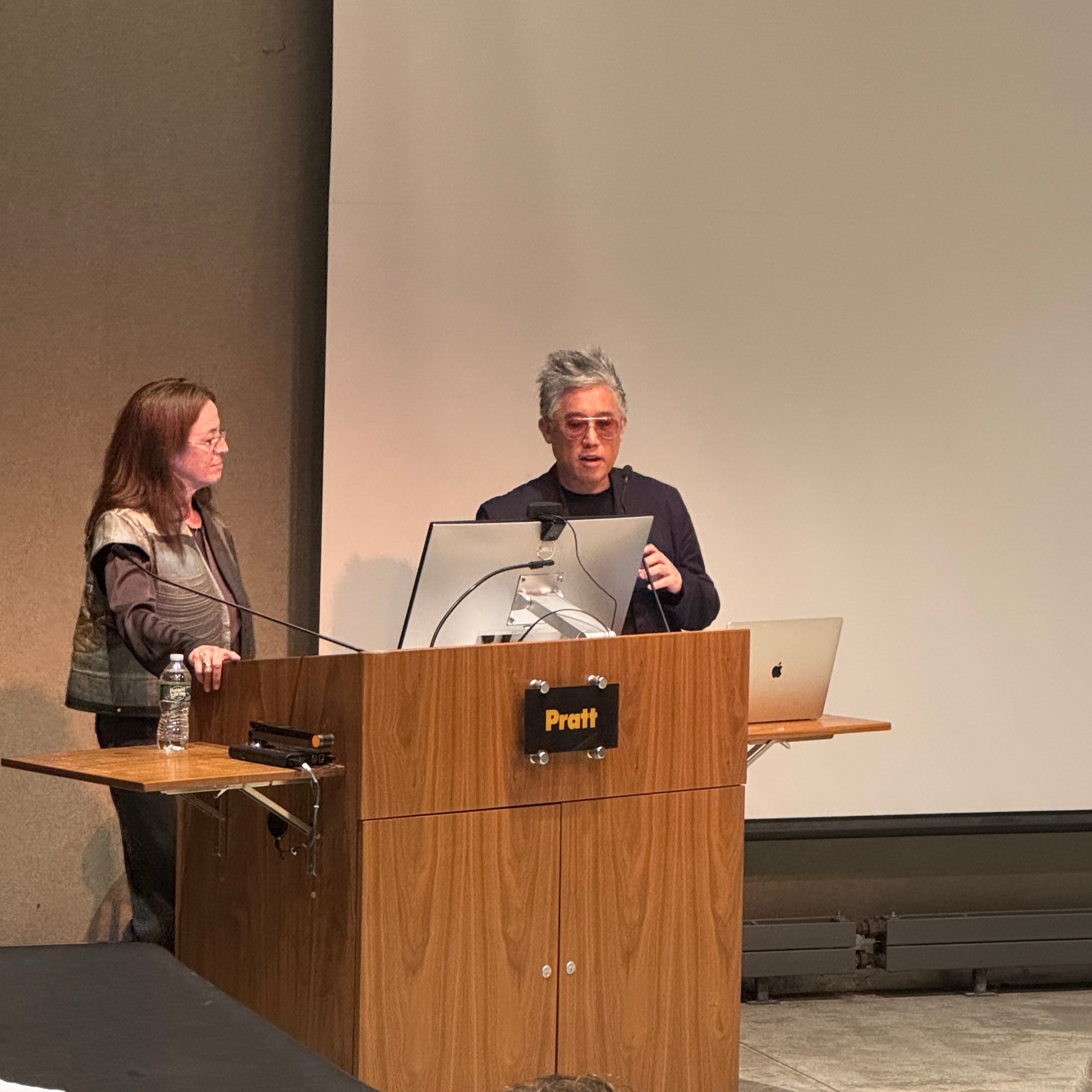 Two speakers standing at a Pratt Institute podium during a lecture. They appear to be presenting slides projected behind them, engaging the audience in a professional setting.
