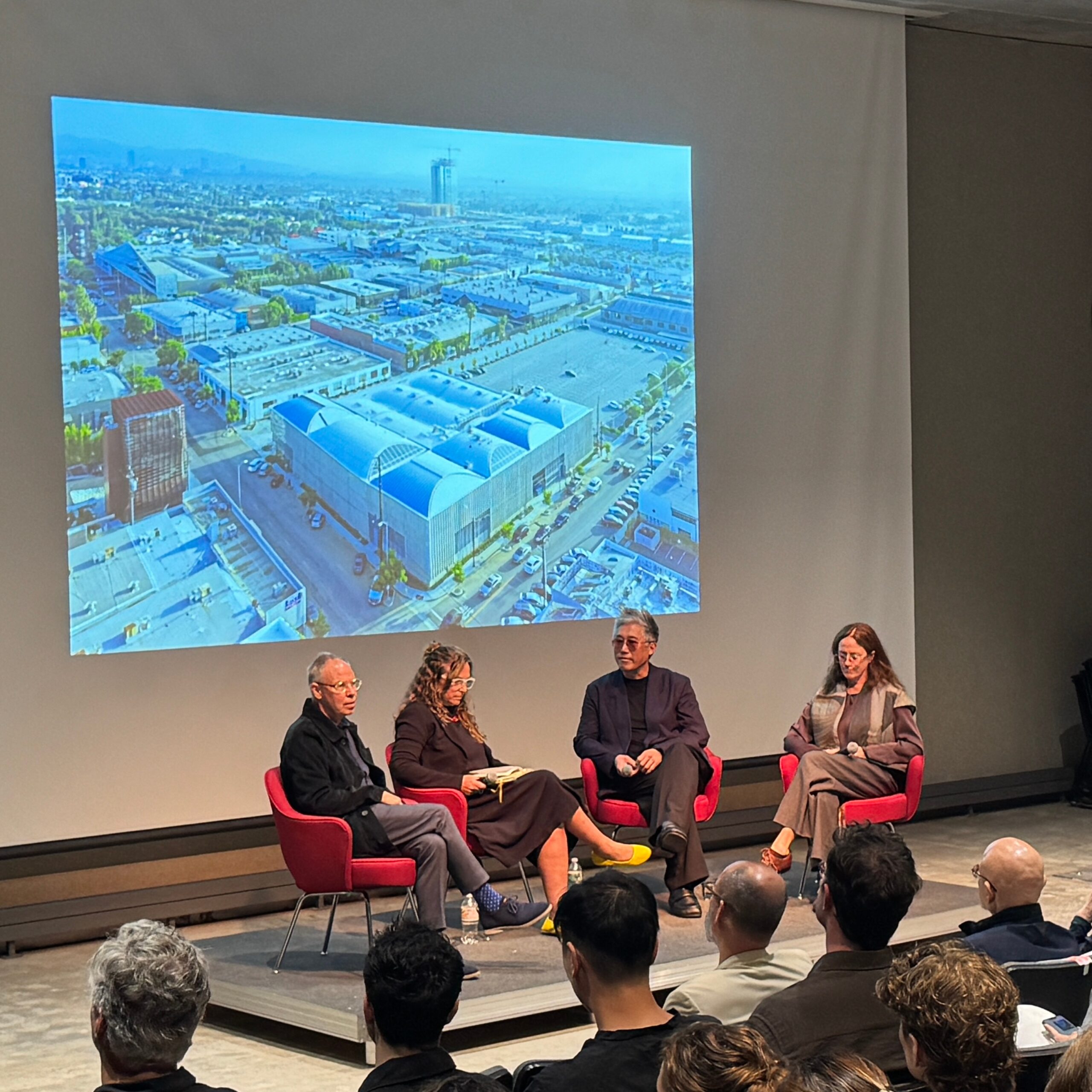 A panel discussion at Pratt Institute featuring four speakers seated on red chairs in front of a large projection of an aerial cityscape. The audience listens attentively to the conversation.