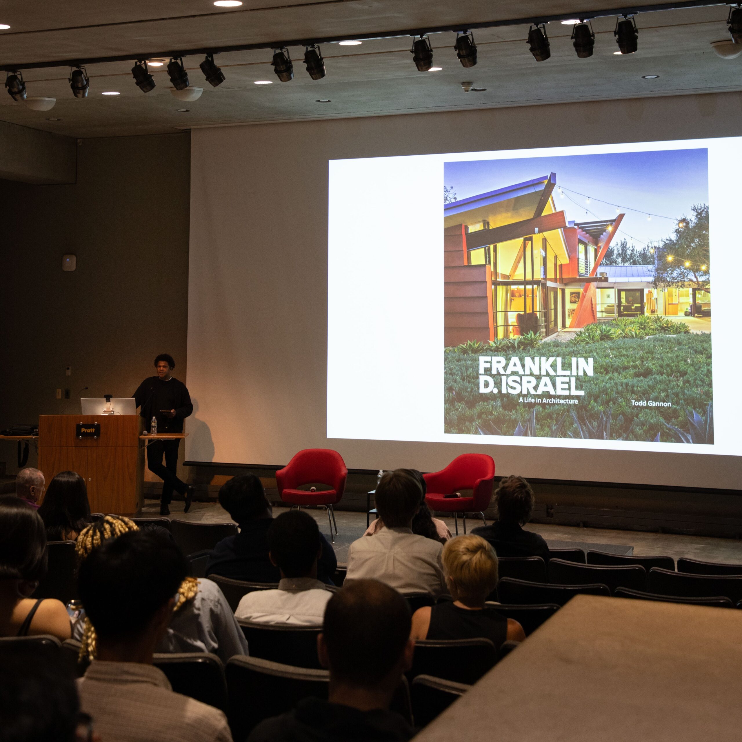 A speaker stands at the Pratt Institute podium giving a lecture, with a large projected image of a modern architectural building labeled “Franklin D. Israel: A Life in Architecture” behind him. Audience members are seated and watching.