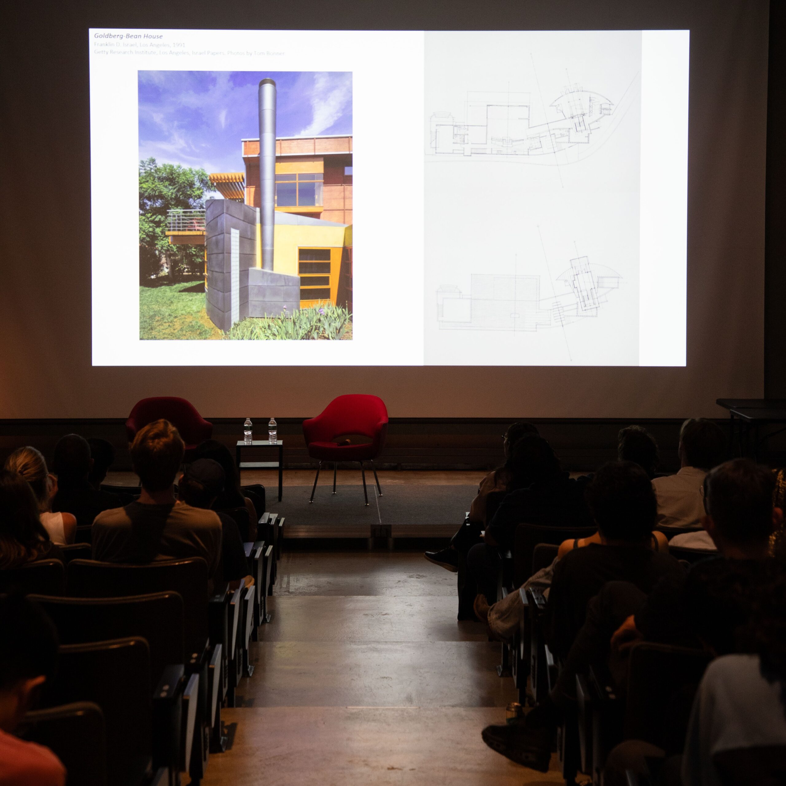 Audience members seated in an auditorium at Pratt Institute watch a presentation showing architectural drawings and a color photograph of a modern house on a large projection screen. Two empty red chairs are positioned on stage.