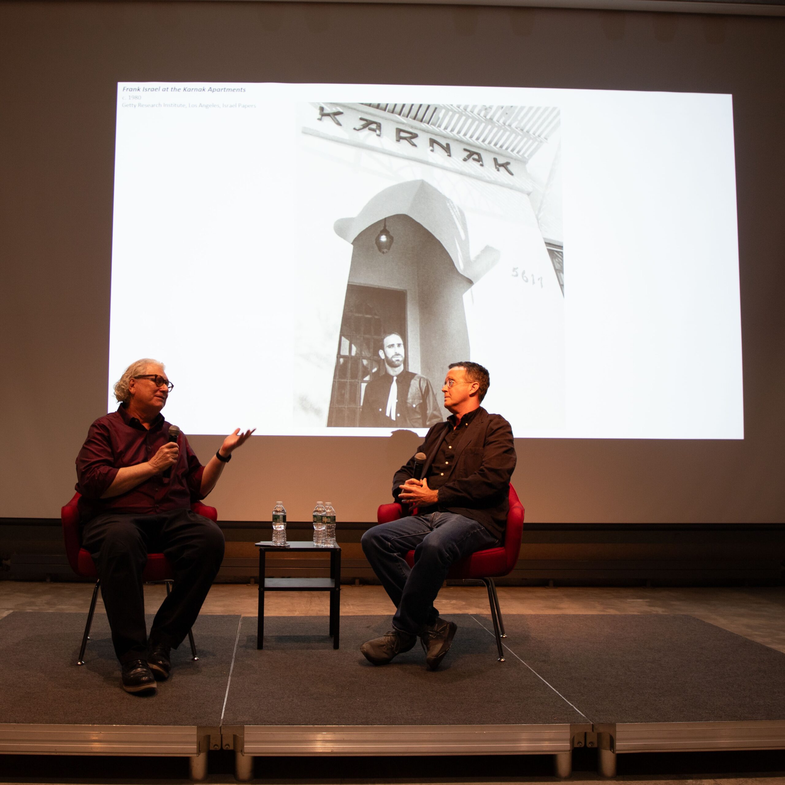 Two individuals sit on stage in red chairs at Pratt Institute, engaging in a discussion before a projected black-and-white image of an architect standing under a sign that reads “KARNAK.” A small table with water bottles sits between them.