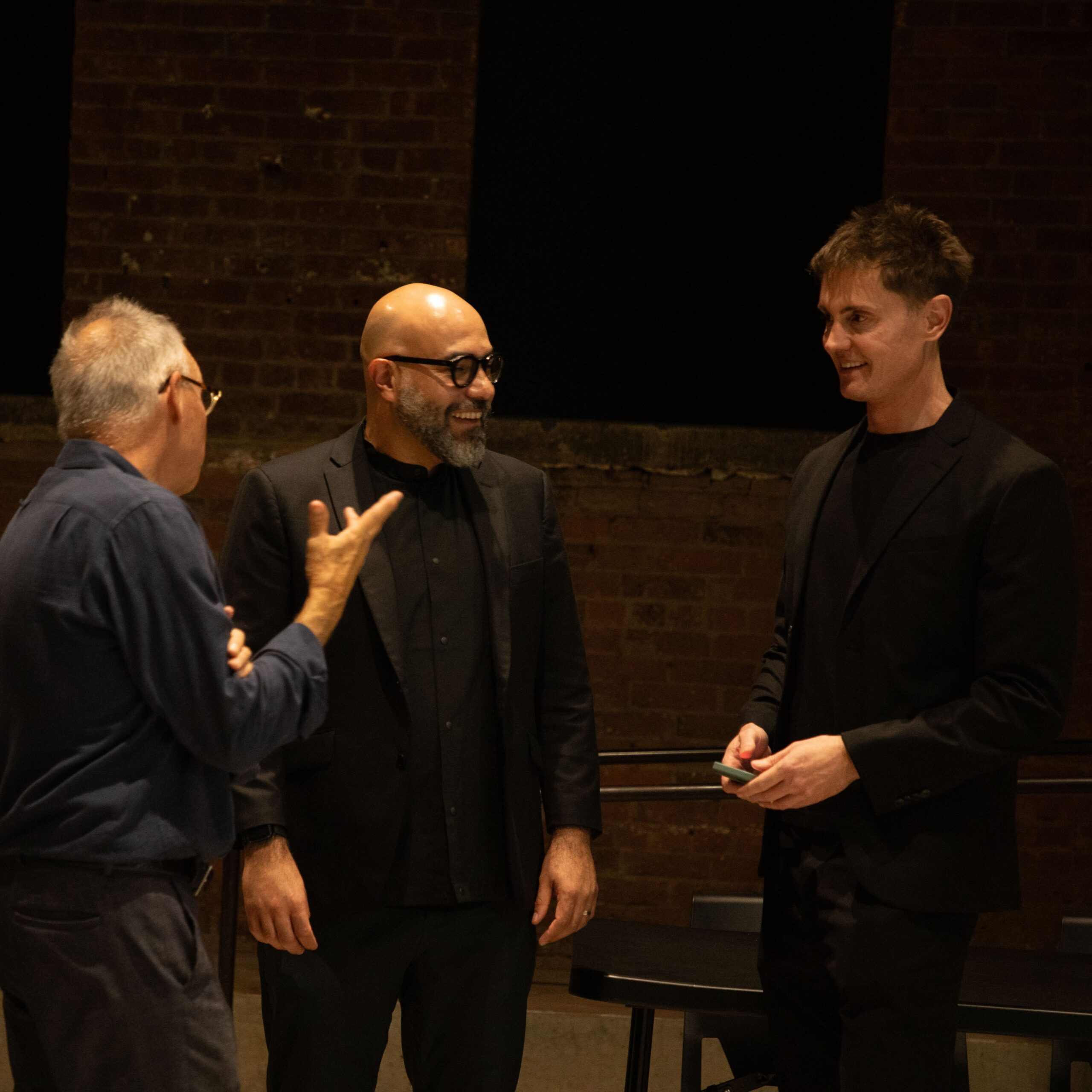 Three individuals standing and conversing in a Pratt Institute auditorium, dressed in formal attire and smiling. The background shows a brick wall and dim lighting typical of a post-event discussion.
