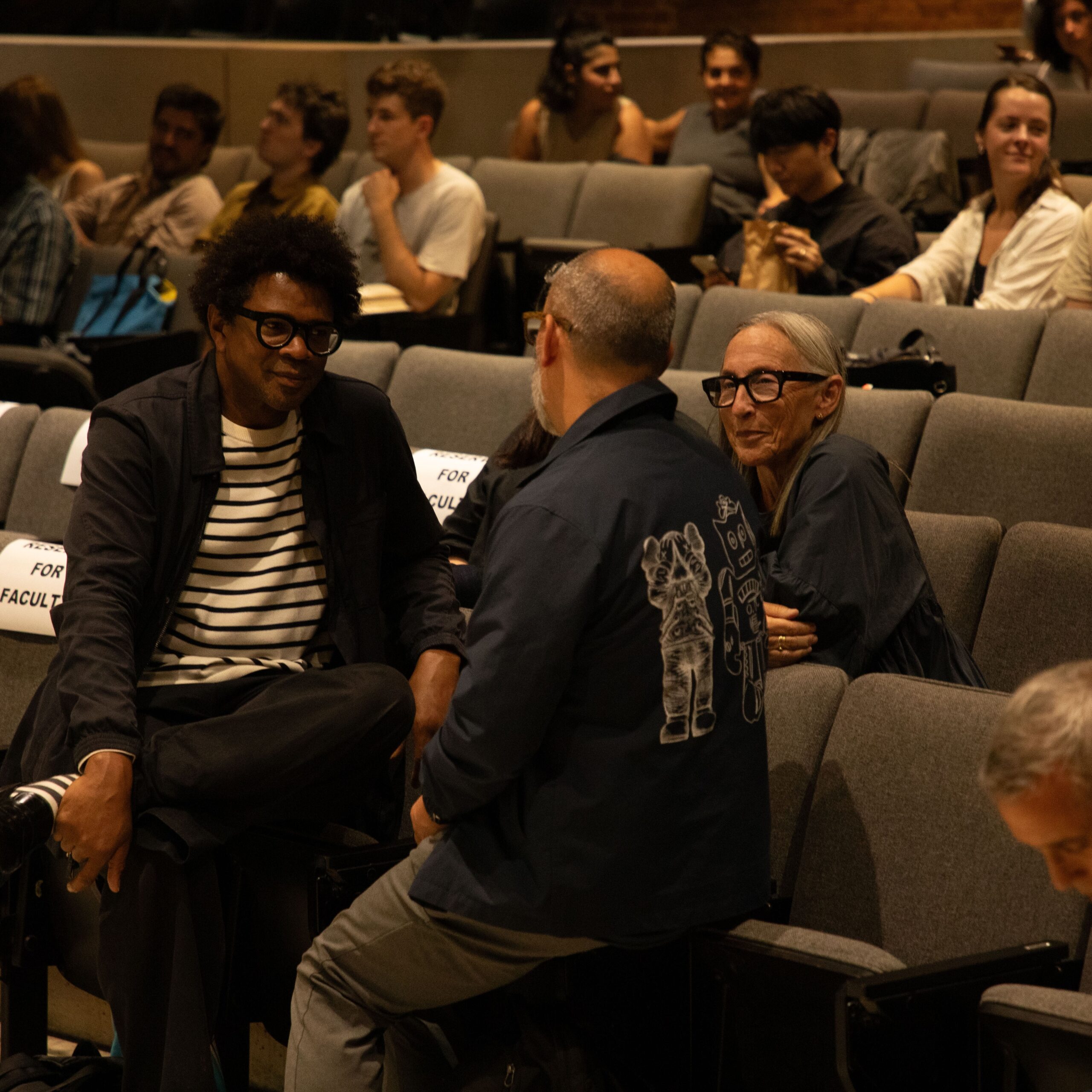Audience members seated in a Pratt Institute auditorium, conversing before a presentation. Three individuals in the foreground are talking and smiling, while others sit and wait in the background.