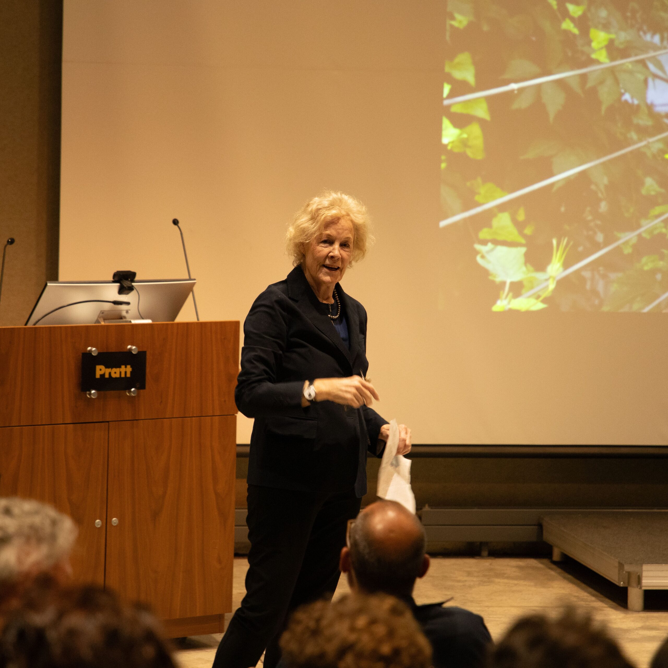 A woman speaking at a Pratt Institute auditorium, standing beside a podium with the Pratt logo. A projected image of green foliage appears behind her as she addresses the audience.