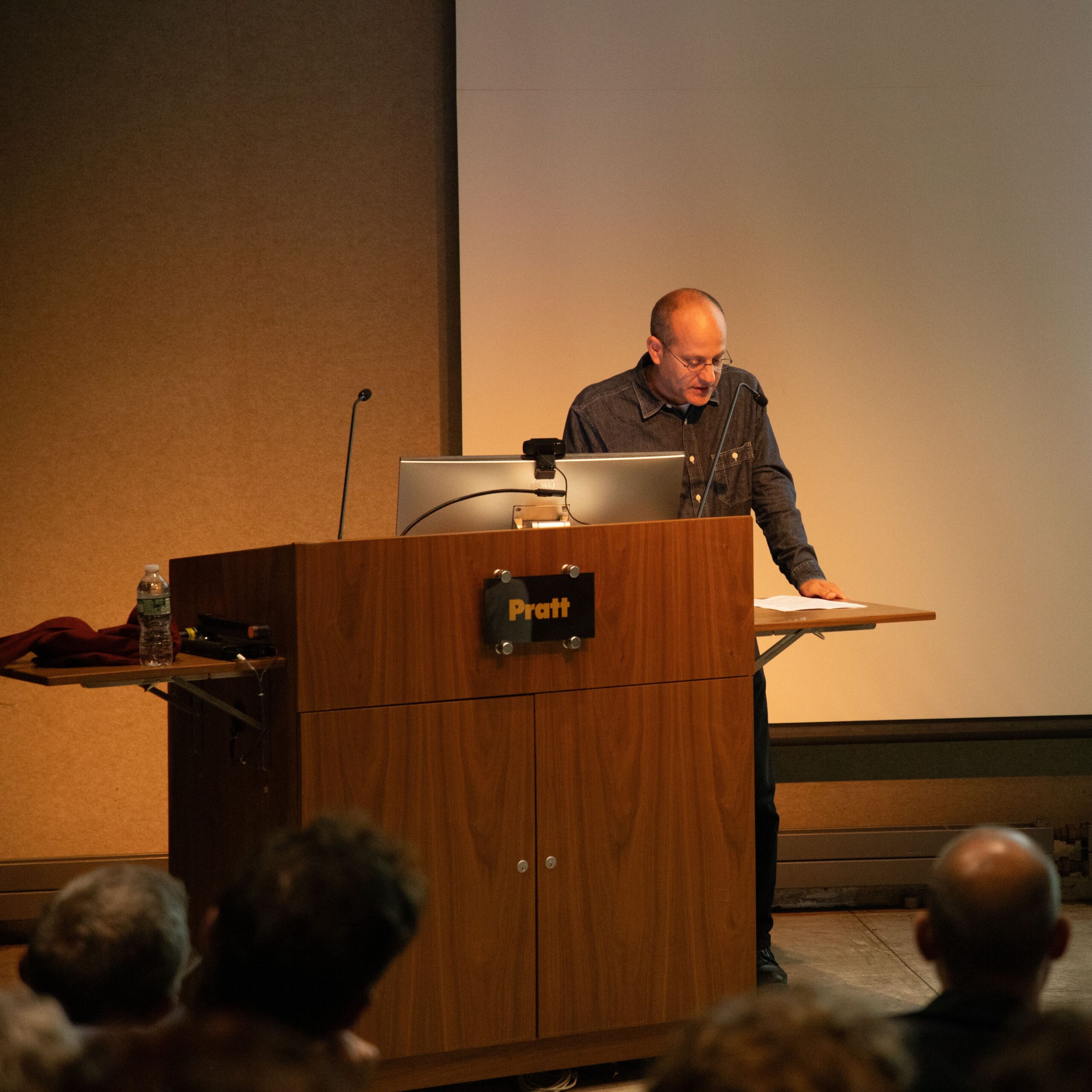 A man standing behind a Pratt Institute podium, reading from notes during a lecture. A projected image is partially visible on the screen behind him as attendees listen attentively.