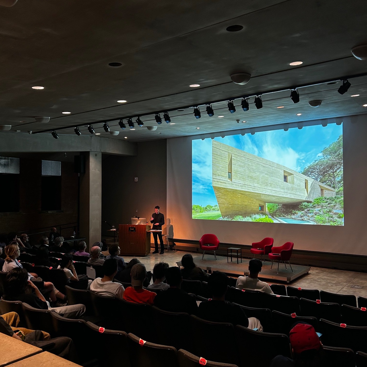 A speaker presents at Pratt Institute’s Higgins Hall auditorium, standing at a podium before a projected image of a modern concrete building under a bright sky. The audience watches attentively from theater-style seating.