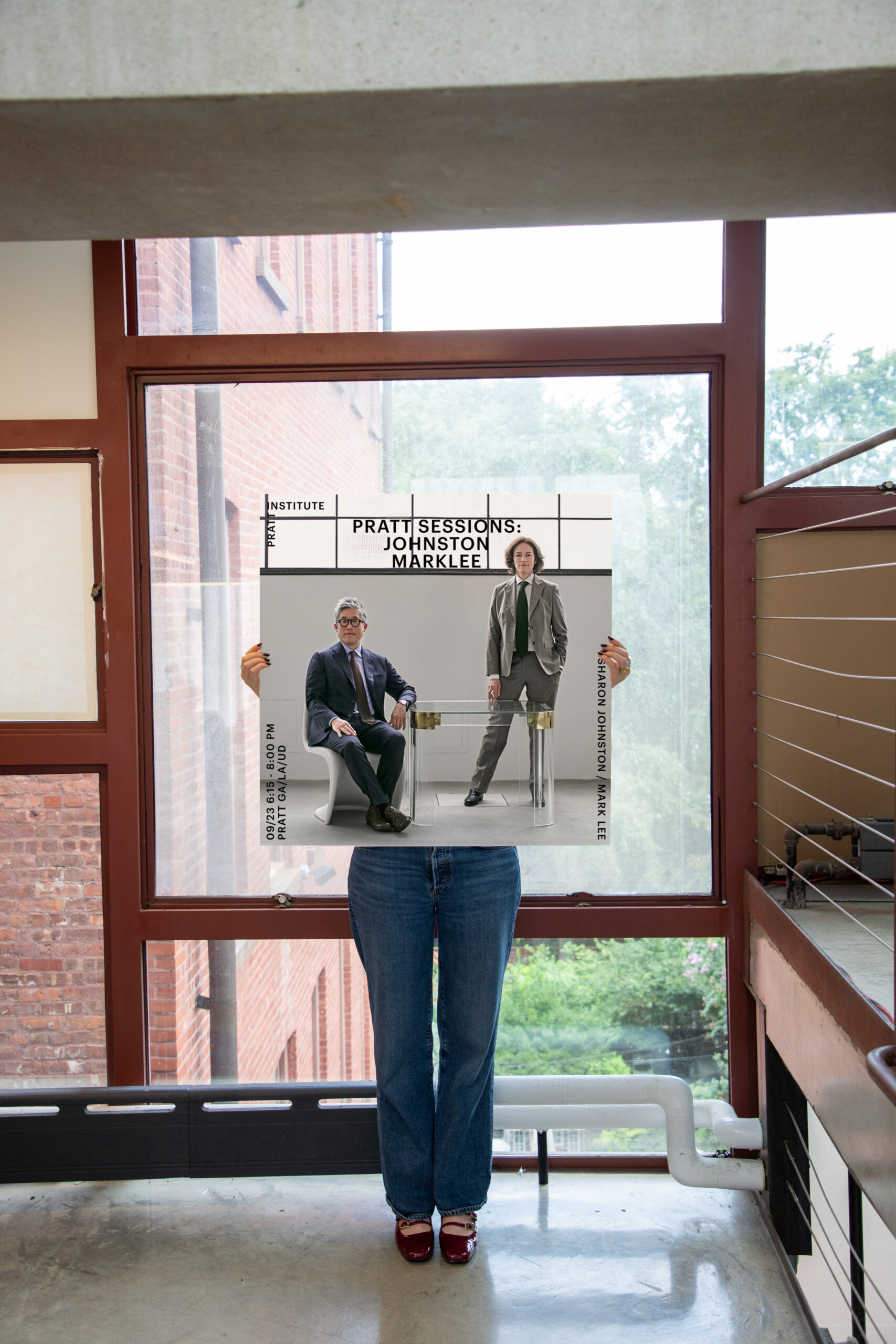 A person standing indoors holding a large poster featuring two individuals—one seated, one standing—promoting the “Pratt Sessions: Johnston Marklee” event. The poster includes event details for a Pratt GA/LA/UD lecture.